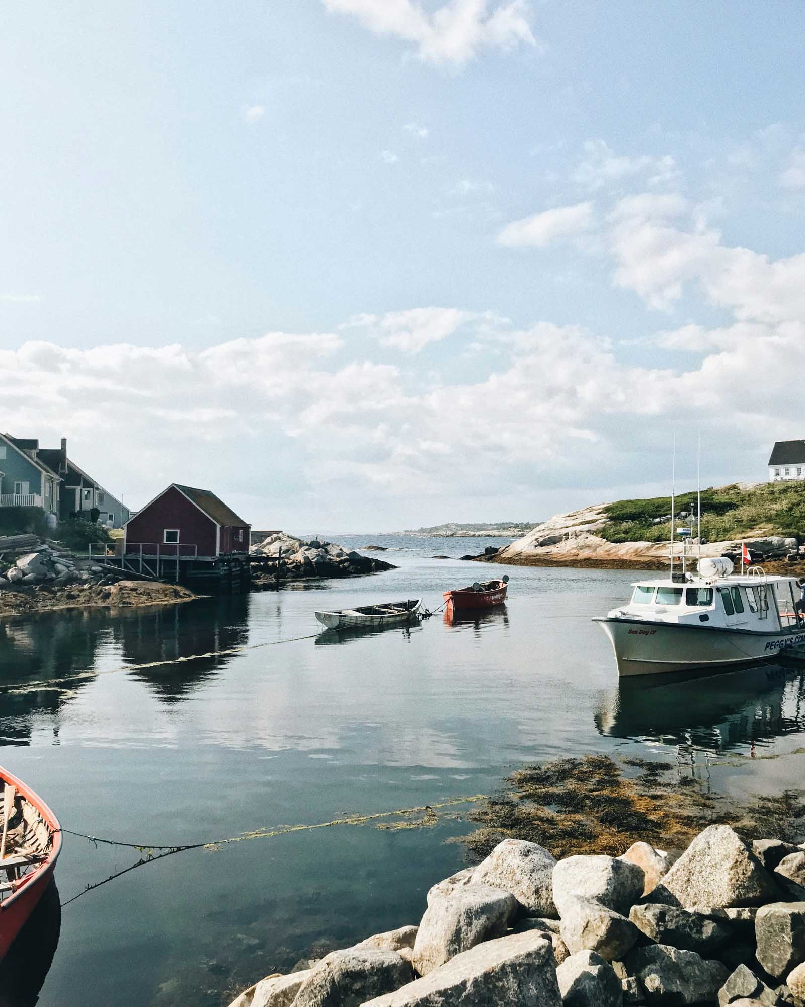 A serene coastal fishing village with traditional wooden buildings, moored fishing boats, and rocky shoreline under a partly cloudy sky. The calm harbor waters reflect the picturesque New England or Maritime charm of this quiet seaside settlement.