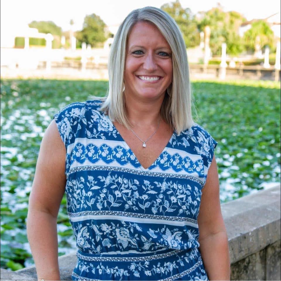 Portrait at Agricultural Farm Setting A smiling woman with blonde hair wearing a blue and white patterned sleeveless top poses for a portrait in front of an agricultural field with blooming crops and farm structures visible in the background.