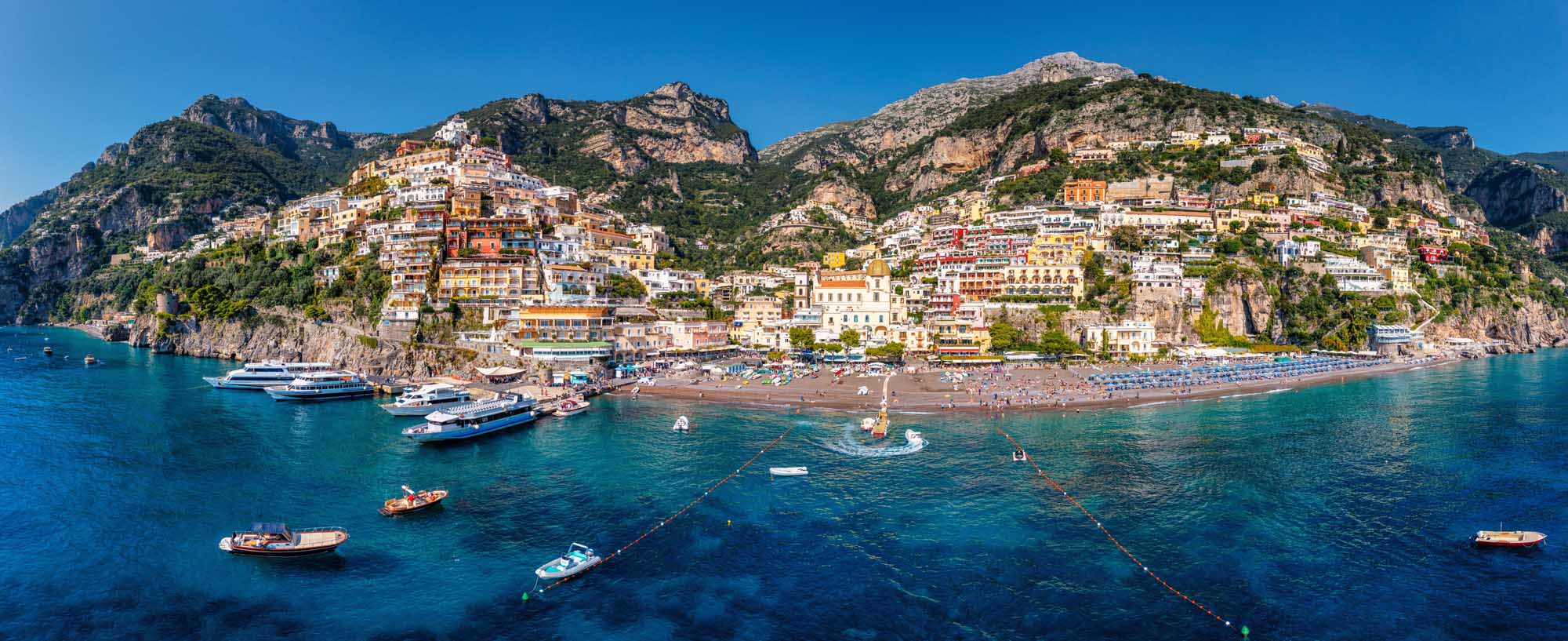 An aerial panoramic view of Positano, a picturesque cliffside village perched dramatically on the Amalfi Coast in Italy. The colorful cascading buildings descend steeply toward a sandy beach and turquoise Mediterranean waters dotted with boats, showcasing the iconic architecture and stunning coastal landscape characteristic of Campania.