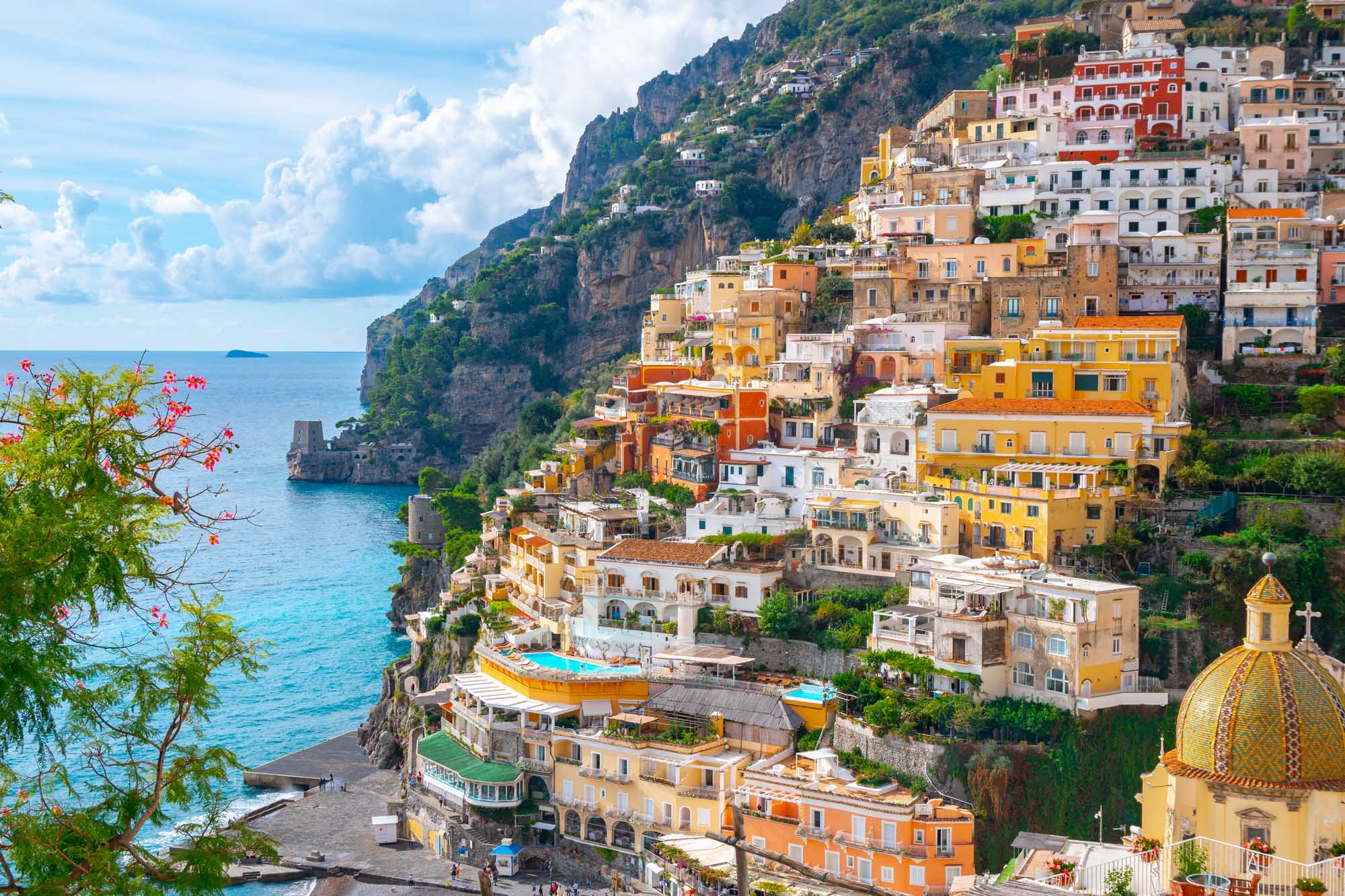 A stunning coastal vista of Positano, Italy, showcasing colorful pastel-hued buildings cascading down steep cliffs to the crystalline Mediterranean Sea. The iconic yellow-domed church (Chiesa di Santa Maria Assunta) is visible on the right, with vibrant yellow, orange, pink, and white buildings stacked vertically along the dramatic rocky coastline, epitomizing the picturesque charm of the Amalfi Coast.