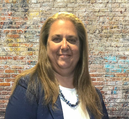 Professional Portrait Against Brick Wall A professional headshot of a woman with long brown hair wearing a dark navy blazer over a white top, posed against a textured brick wall background with weathered red, orange, and blue paint.