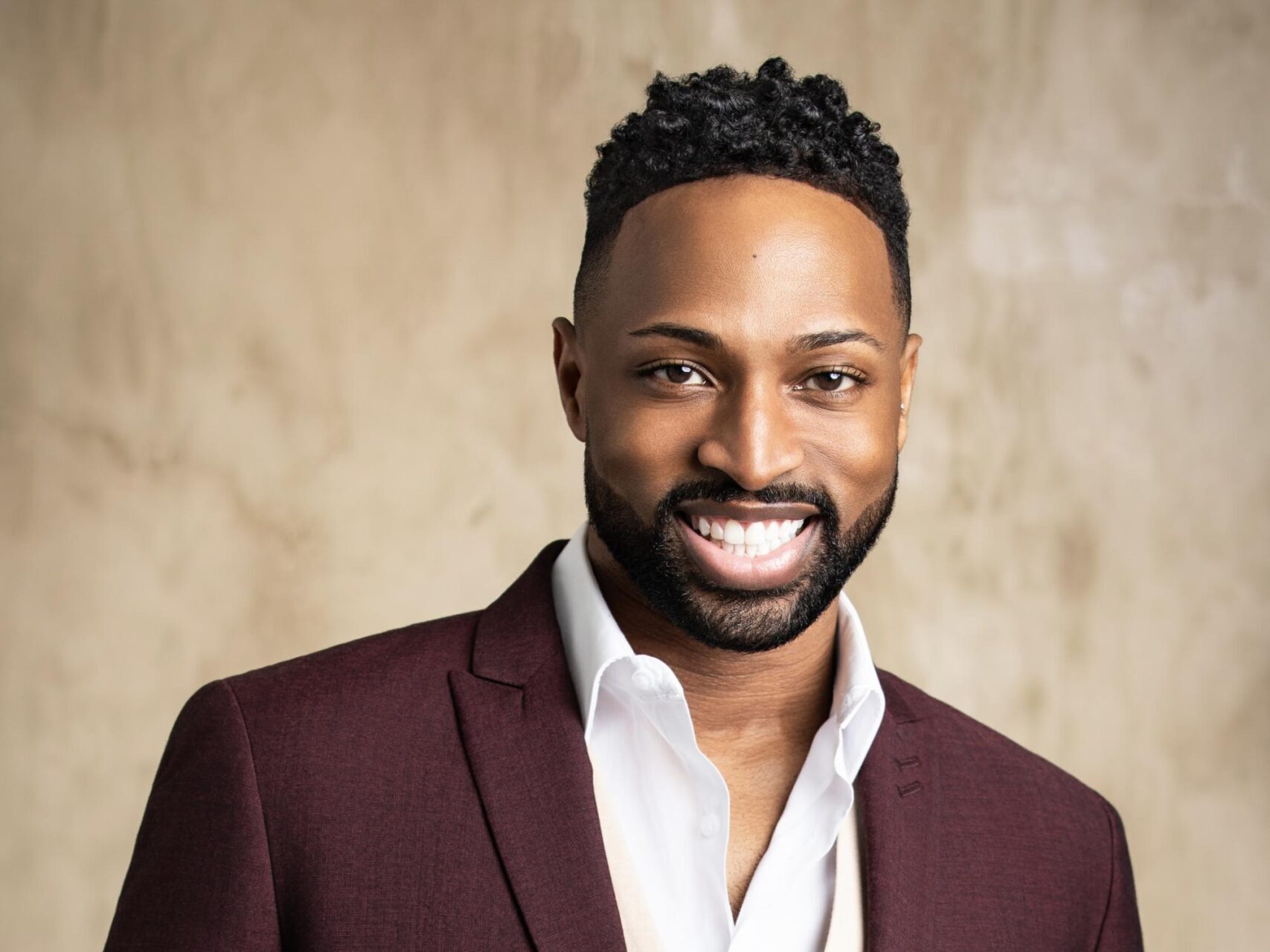 A professional headshot of a man wearing a burgundy blazer over a white shirt, smiling at