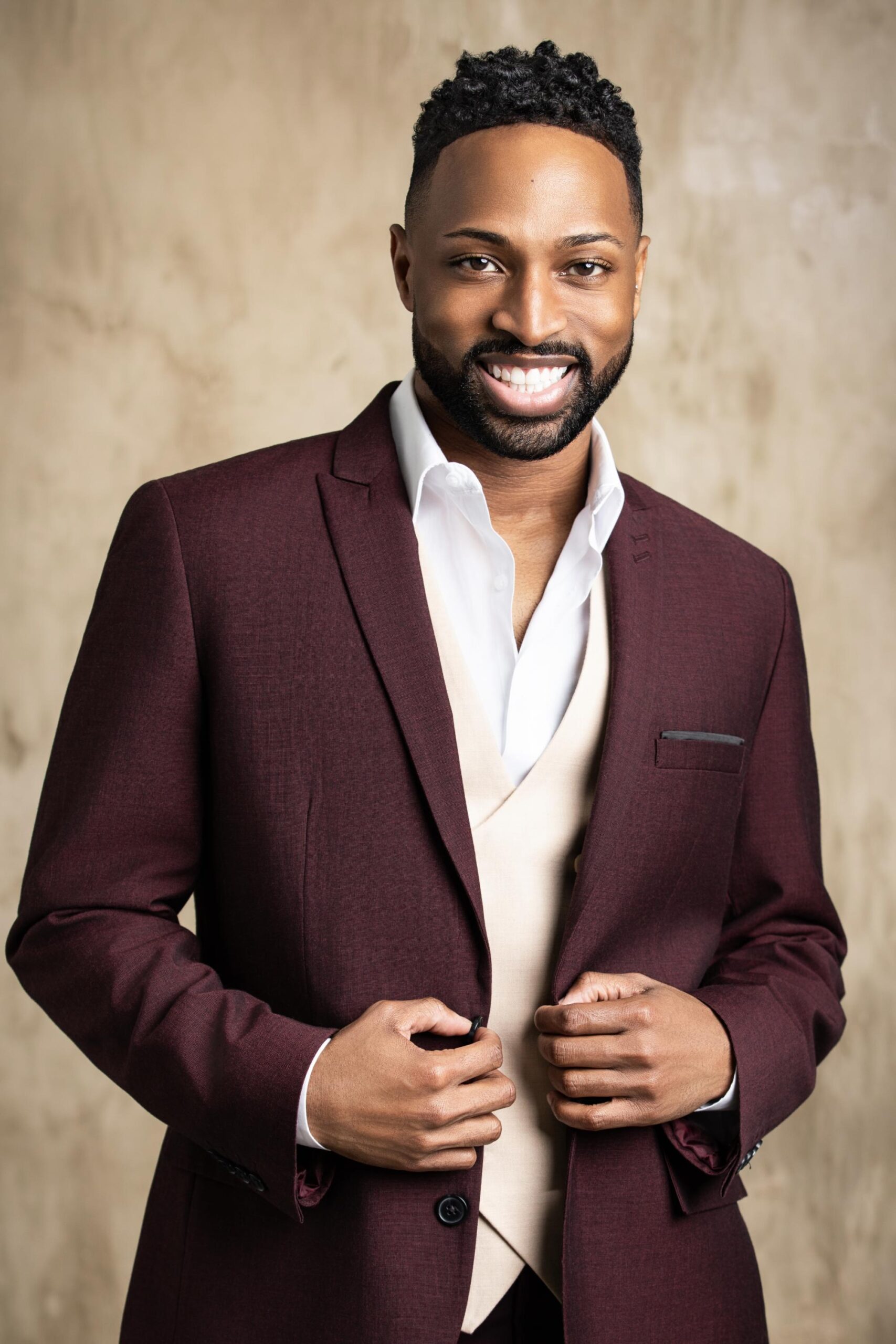 Professional Portrait in Burgundy Blazer A professional headshot of a man wearing a burgundy blazer over a white shirt, smiling at the camera against a neutral beige background. This is a polished corporate or business portrait style photograph.