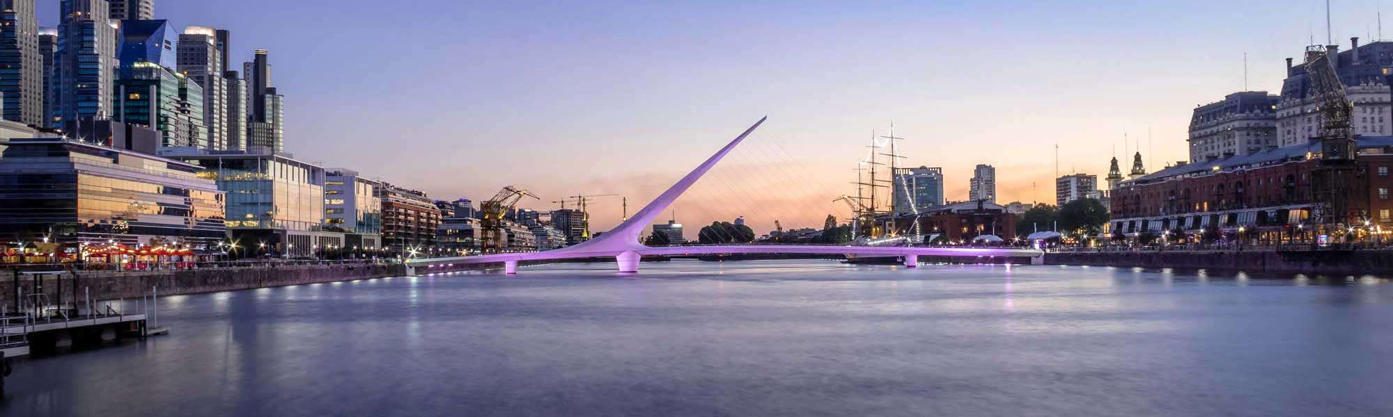 Puente de la Mujer Bridge at Dusk, Buenos Aires A stunning twilight view of the iconic Puente de la Mujer (Woman's Bridge) illuminated in purple lights, spanning a calm waterway in the Puerto Madero neighborhood of Buenos Aires. The modern cable-stayed bridge is framed by contemporary architecture, historic buildings, and tall ships, creating a striking contrast between old and new urban development.