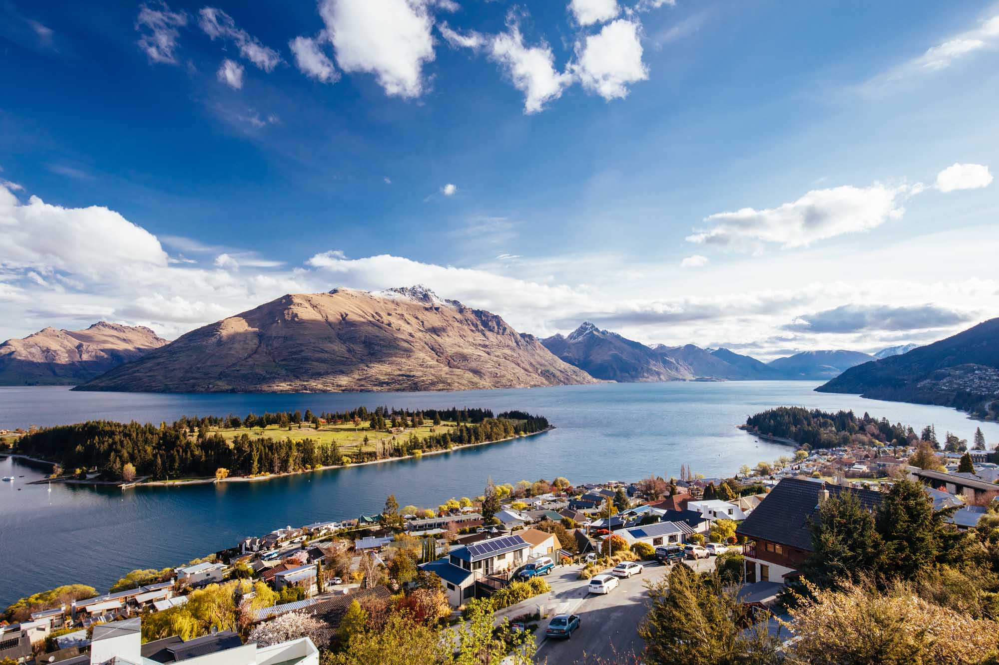 Queenstown Lakeside Town with Mountain Views An aerial view of Queenstown, New Zealand, nestled on the shores of Lake Wakatipu with snow-capped mountains in the background. The scenic alpine landscape features a charming residential community surrounded by forested peninsulas and crystal-clear turquoise waters, with Cecil Peak prominently visible across the lake under a brilliant blue sky with white clouds.