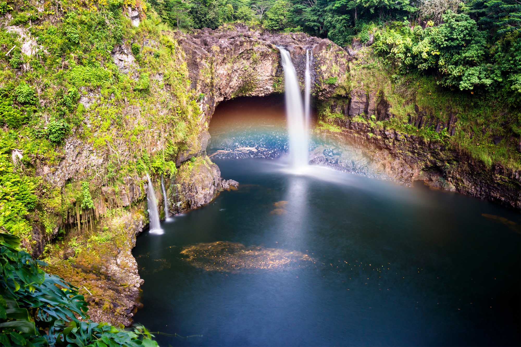 Rainbow Falls Waterfall in Hilo Hawaii A stunning waterfall cascades into a deep natural pool surrounded by lush green vegetation and moss-covered basalt cliffs in Hilo, Hawaii. The pristine turquoise waters and verdant landscape create a serene tropical paradise, with the waterfall's misty spray catching sunlight to create subtle rainbow effects.