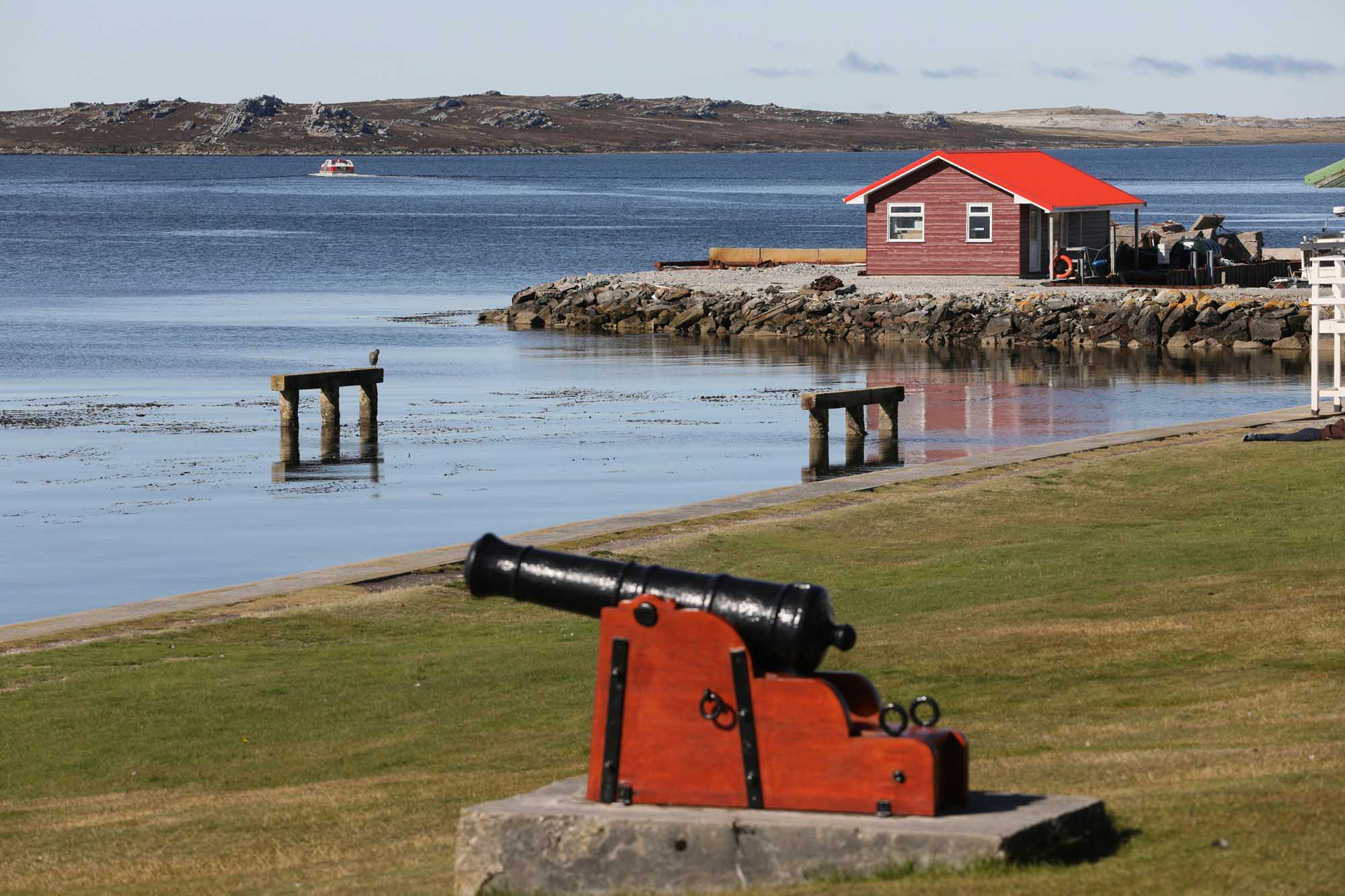 Red Boathouse and Cannon on Arctic Coastline A picturesque Arctic coastal settlement featuring a distinctive red-roofed boathouse perched on a rocky peninsula, with a red cannon in the foreground on manicured green grass. The calm blue waters, weathered wooden structures, and barren hillsides in the background are characteristic of remote Greenlandic or Faroese communities.