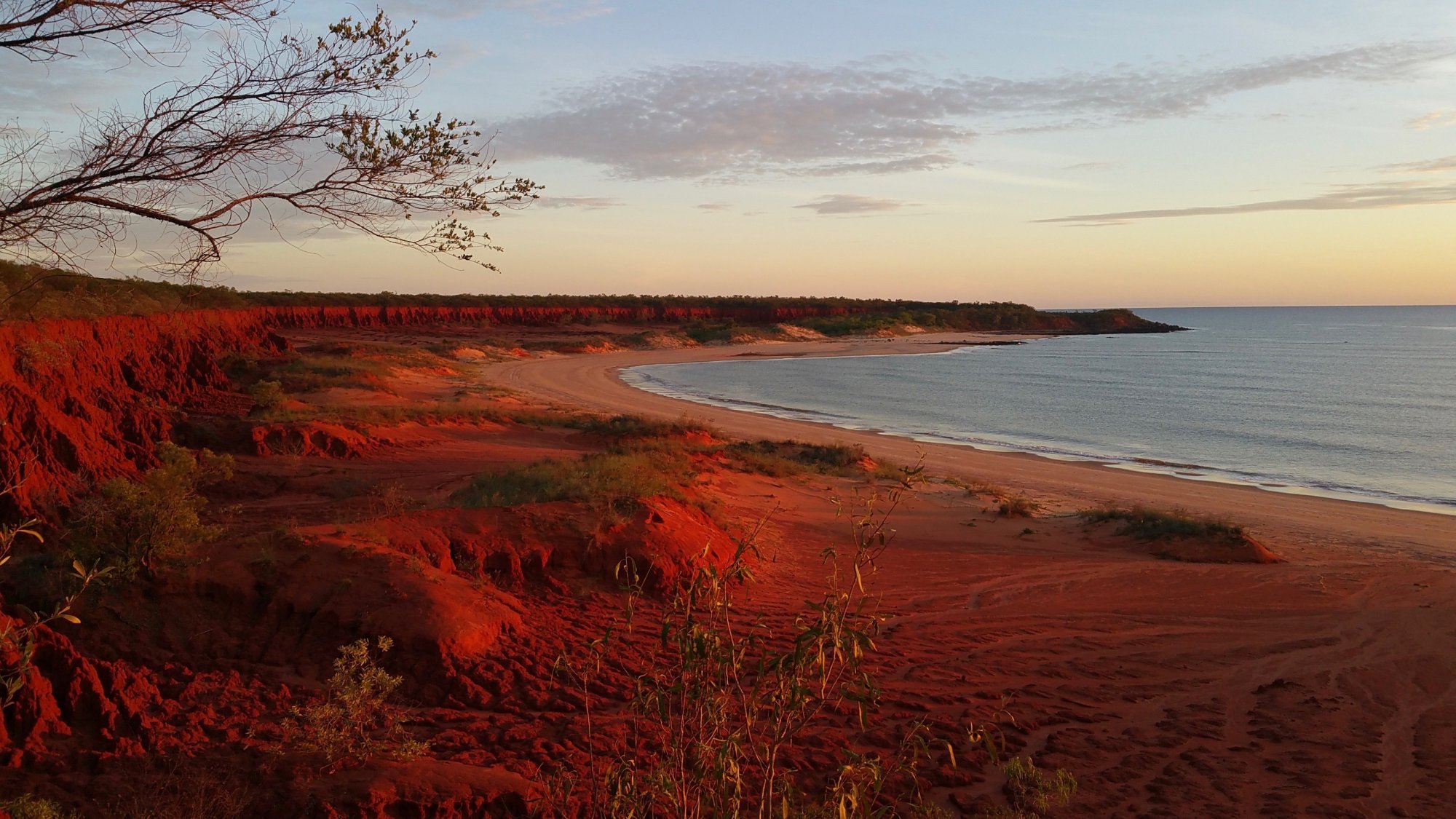Red Cliffs Coastal Beach at Sunset A stunning coastal landscape featuring dramatic red sandstone cliffs overlooking a curved sandy beach and calm waters at dusk. The warm golden light of sunset illuminates the distinctive rust-colored earth formations, creating a striking contrast with the blue-grey ocean and pale sky beyond.