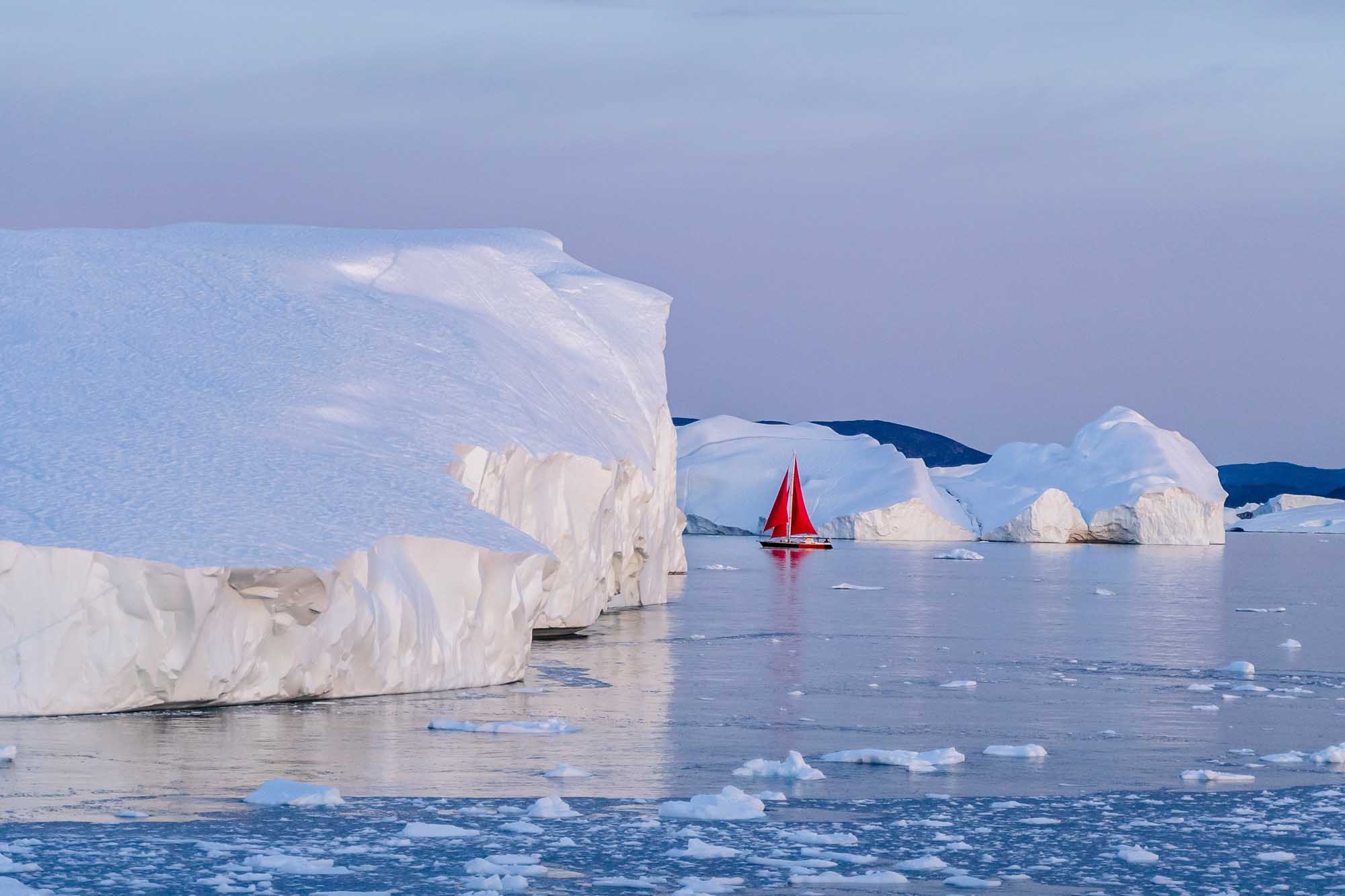 Red Sailboat Among Icebergs in Arctic Waters A striking red-sailed vessel navigates through a pristine Arctic landscape dotted with massive white icebergs. The scene captures the contrast between the vivid crimson sail and the pale blue waters and towering ice formations, showcasing exploration in one of Earth's most remote and dramatic polar regions.
