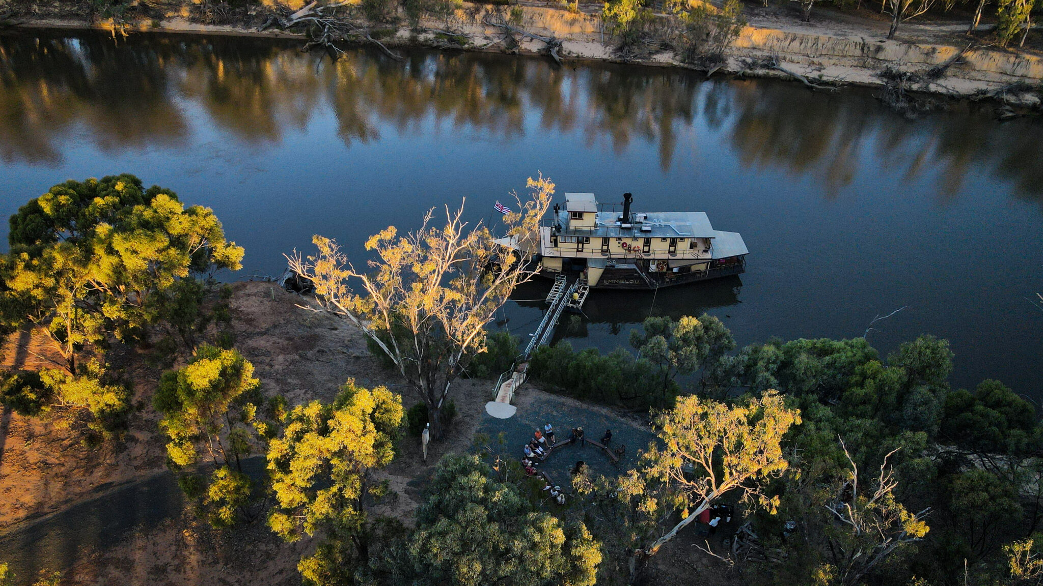 River Cruise Vessel at Scenic Landing An aerial view of a multi-deck river cruise ship moored at a sandy riverbank landing with passengers gathered on shore surrounded by golden-lit eucalyptus trees. The vessel features a distinctive white and cream livery with multiple deck levels, connected to the landing via a gangway, on a calm, mirror-like river with dramatic cliff faces visible in the background.