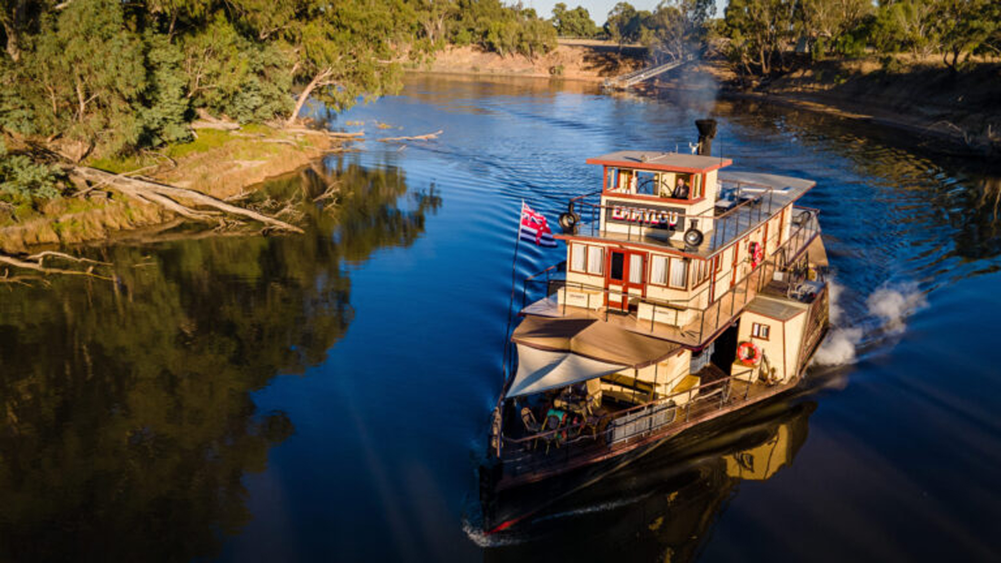 Riverboat Cruise on Australian Inland Waterway A multi-deck paddle steamer or riverboat navigates a serene inland river lined with native eucalyptus trees and lush vegetation. The vessel features distinctive orange and cream-colored superstructure with multiple decks, railings, and a flag, representing a classic Australian river cruise experience in what appears to be a regional waterway.