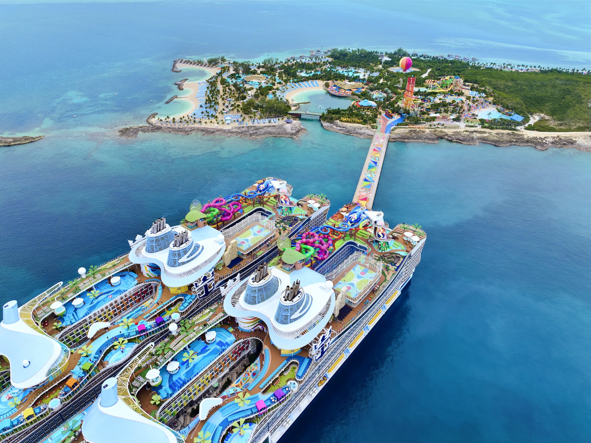 An aerial view of multiple Royal Caribbean cruise ships docked at Perfect Day at CocoCay, a private island destination in the Bahamas. The image showcases the resort's vibrant water park, colorful slides, beach areas, and lush landscaping, with several massive cruise vessels moored alongside a long causeway connecting to the island's attractions.
