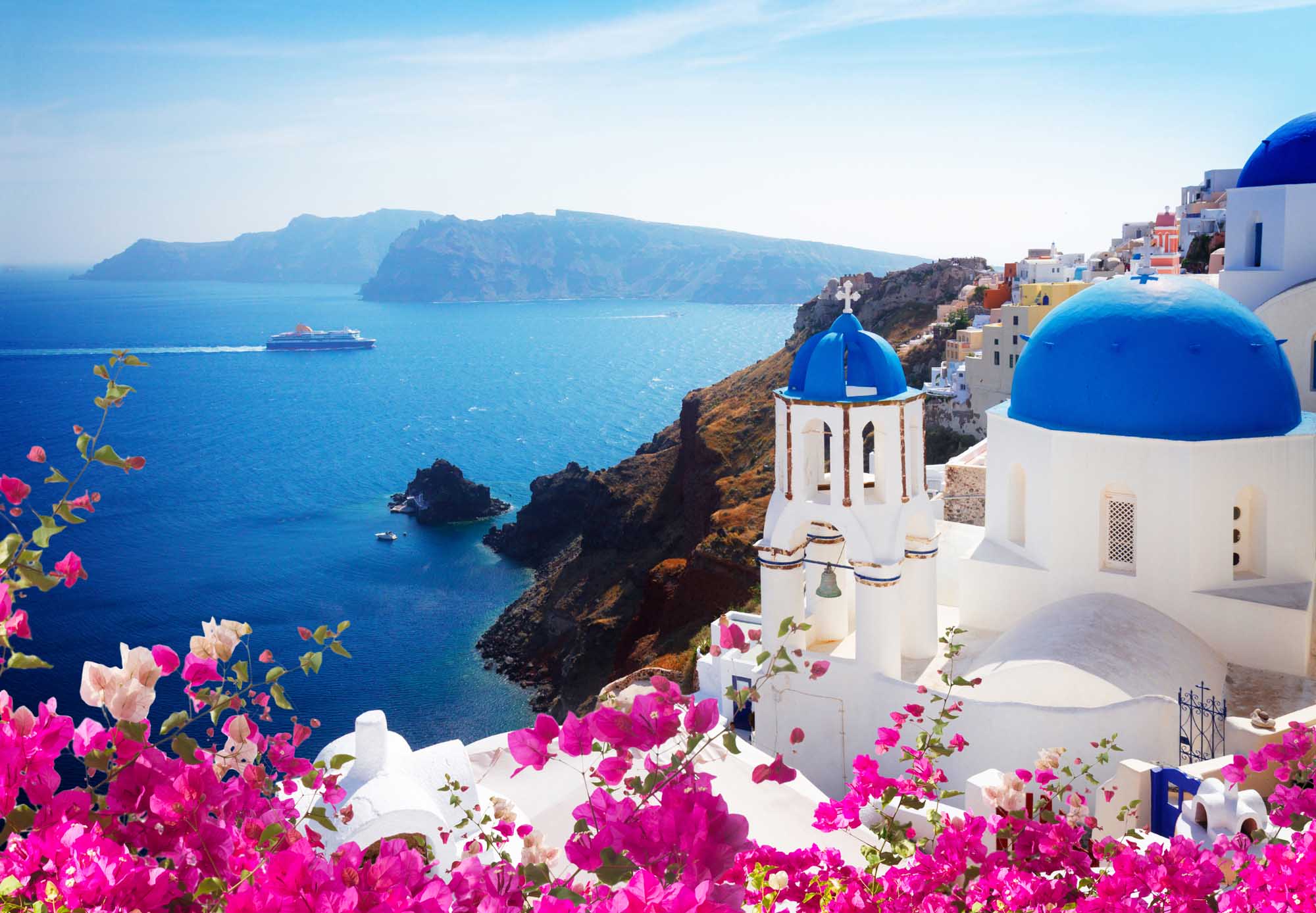 A picturesque view of Oia village in Santorini, Greece, featuring iconic white-washed buildings with brilliant blue-domed churches overlooking the deep blue Aegean Sea. Vibrant magenta bougainvillea flowers frame the foreground, with a cruise ship visible in the distance and the volcanic caldera landscape providing a dramatic backdrop.