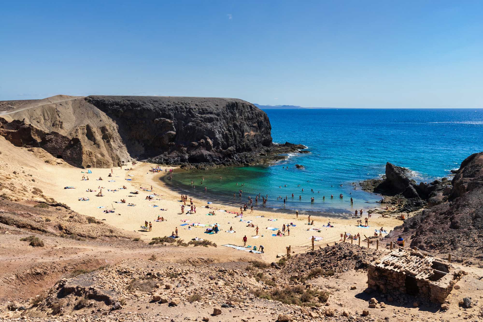 A pristine sandy beach nestled beneath towering dark volcanic cliffs, featuring crystal-clear turquoise waters and numerous sunbathers enjoying the Mediterranean summer. The sheltered cove is surrounded by rugged rock formations, creating a dramatic and picturesque coastal landscape.