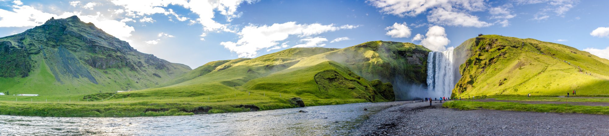 A stunning panoramic view of Seljalandsfoss waterfall cascading down moss-covered cliffs in southern Iceland, with lush green mountains, a glacial river in the foreground, and visitors exploring the scenic landscape under a partly cloudy sky.