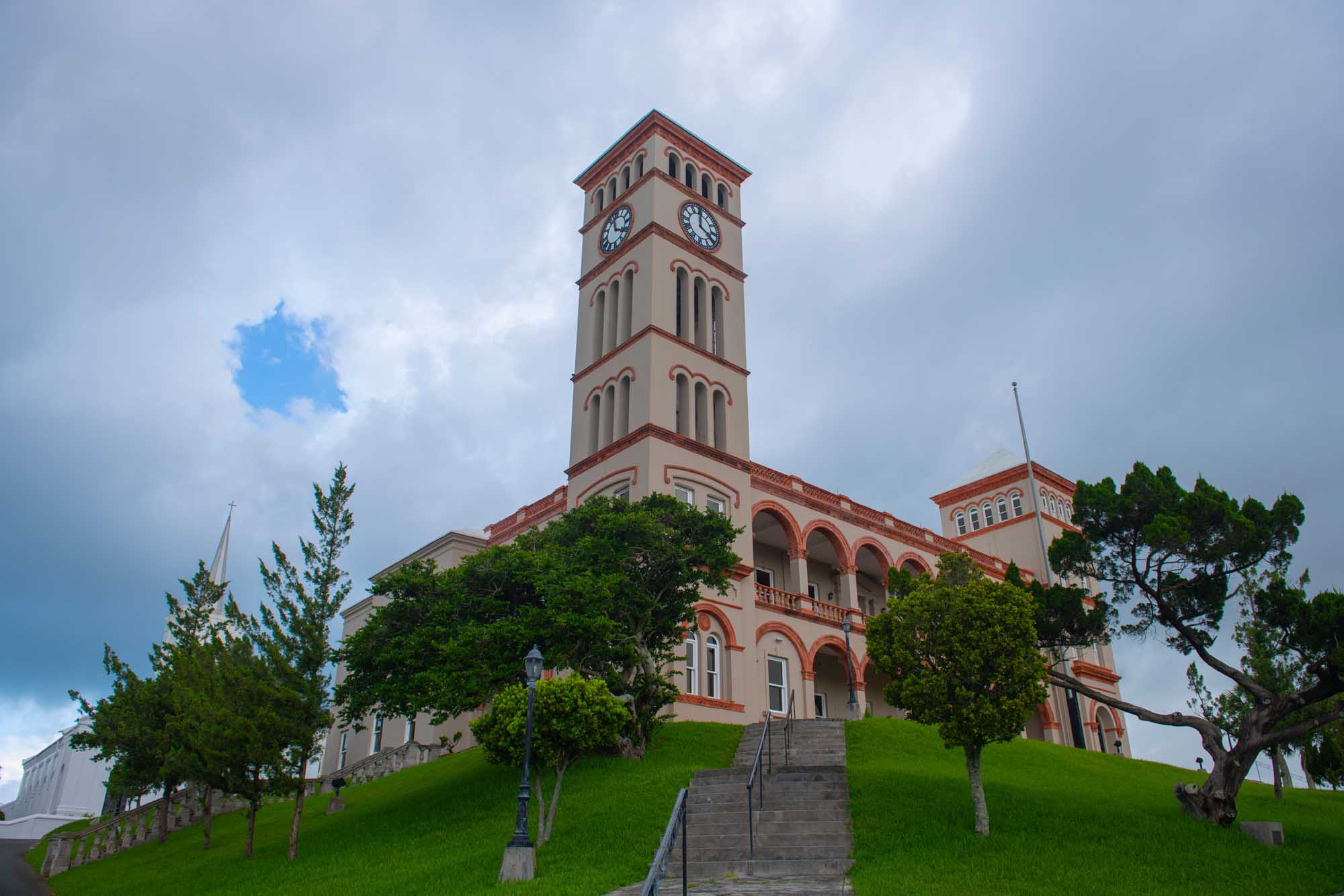 A distinctive pink and cream-colored colonial government building featuring a prominent clock tower, situated on a manicured green hillside in Hamilton's city center. The Sessions House serves as the home of the House of Assembly and Supreme Court of Bermuda, showcasing classic Bermudian architecture with arched windows and ornate details.
