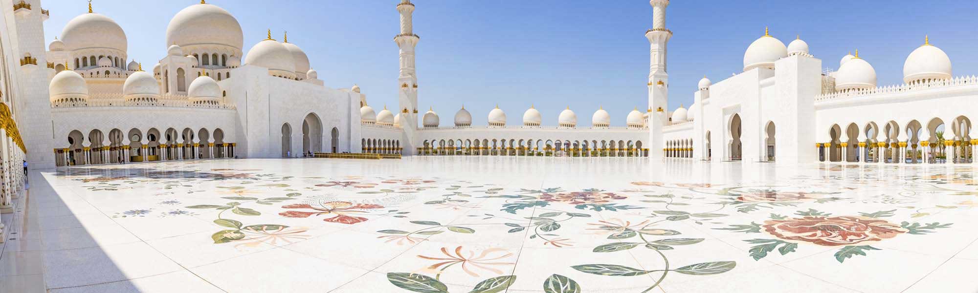 The stunning courtyard of Sheikh Zayed Grand Mosque in Abu Dhabi, featuring pristine white marble flooring adorned with intricate floral inlay patterns in colorful stones. The mosque's iconic white domes, minarets, and ornate golden architectural details are visible under a clear blue sky, creating a serene and majestic Islamic architectural landscape.