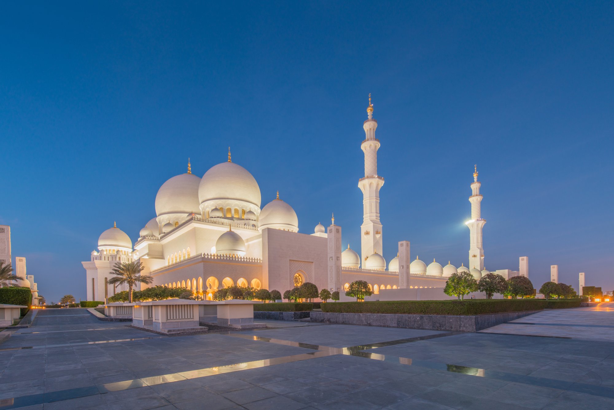 Sheikh Zayed Grand Mosque at Twilight The iconic Sheikh Zayed Grand Mosque illuminated at dusk against a deep blue sky, featuring pristine white marble architecture with multiple domes and minarets, reflected in the still water of the courtyard plaza. The mosque's stunning Islamic design and symmetrical layout showcase the architectural grandeur of Abu Dhabi's most significant religious landmark.