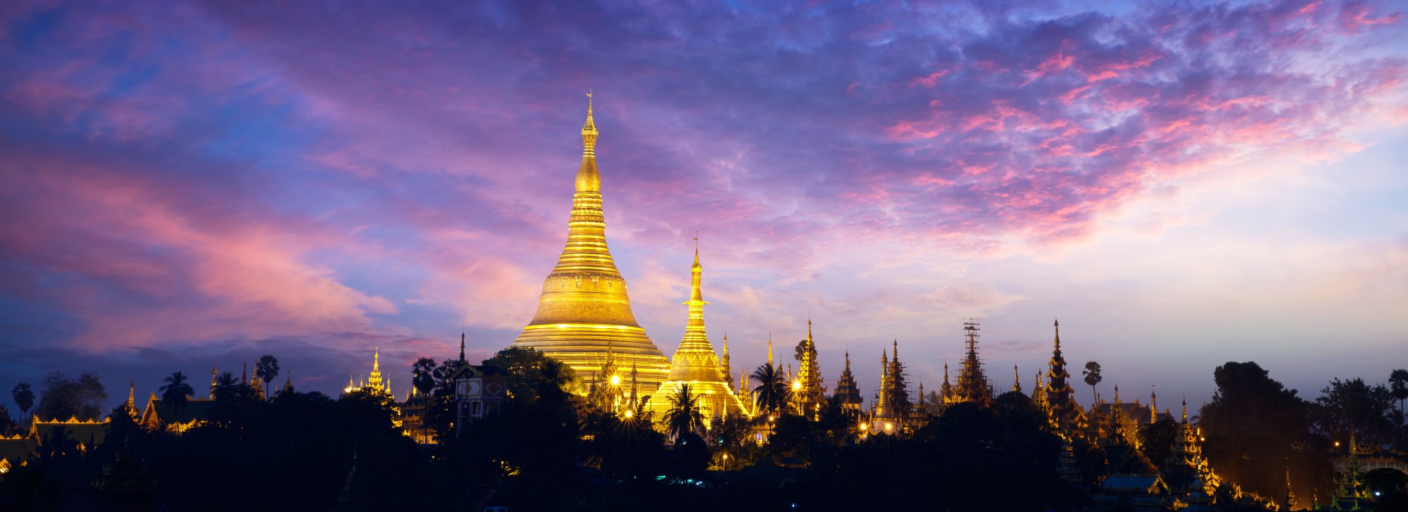 Shwedagon Pagoda at Twilight The iconic golden Shwedagon Pagoda illuminated against a dramatic purple and pink twilight sky, with its distinctive tiered spire rising prominently above the surrounding temple complex and palm trees. The sacred Buddhist monument glows warmly in the evening light, showcasing the intricate architecture of Myanmar's most revered spiritual landmark.