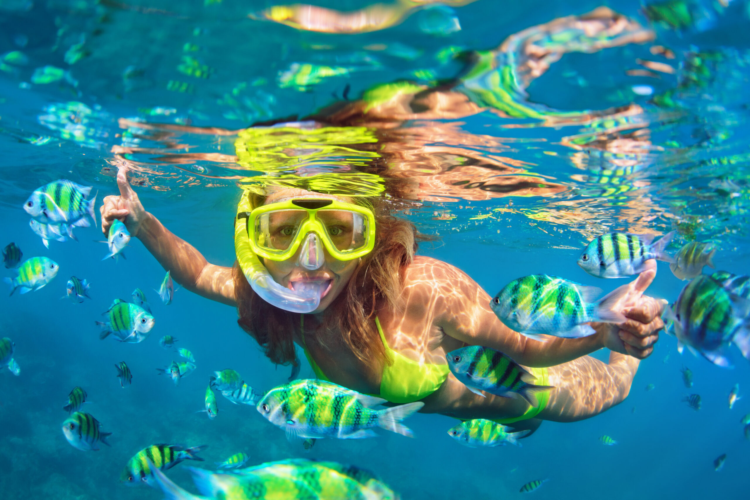 A young woman snorkels in crystal-clear tropical waters wearing a bright yellow-green snorkel mask and matching swimwear, surrounded by vibrant striped tropical fish with yellow and blue markings. She smiles at the camera with her arms outstretched, capturing the joy and wonder of underwater exploration in a pristine marine environment.