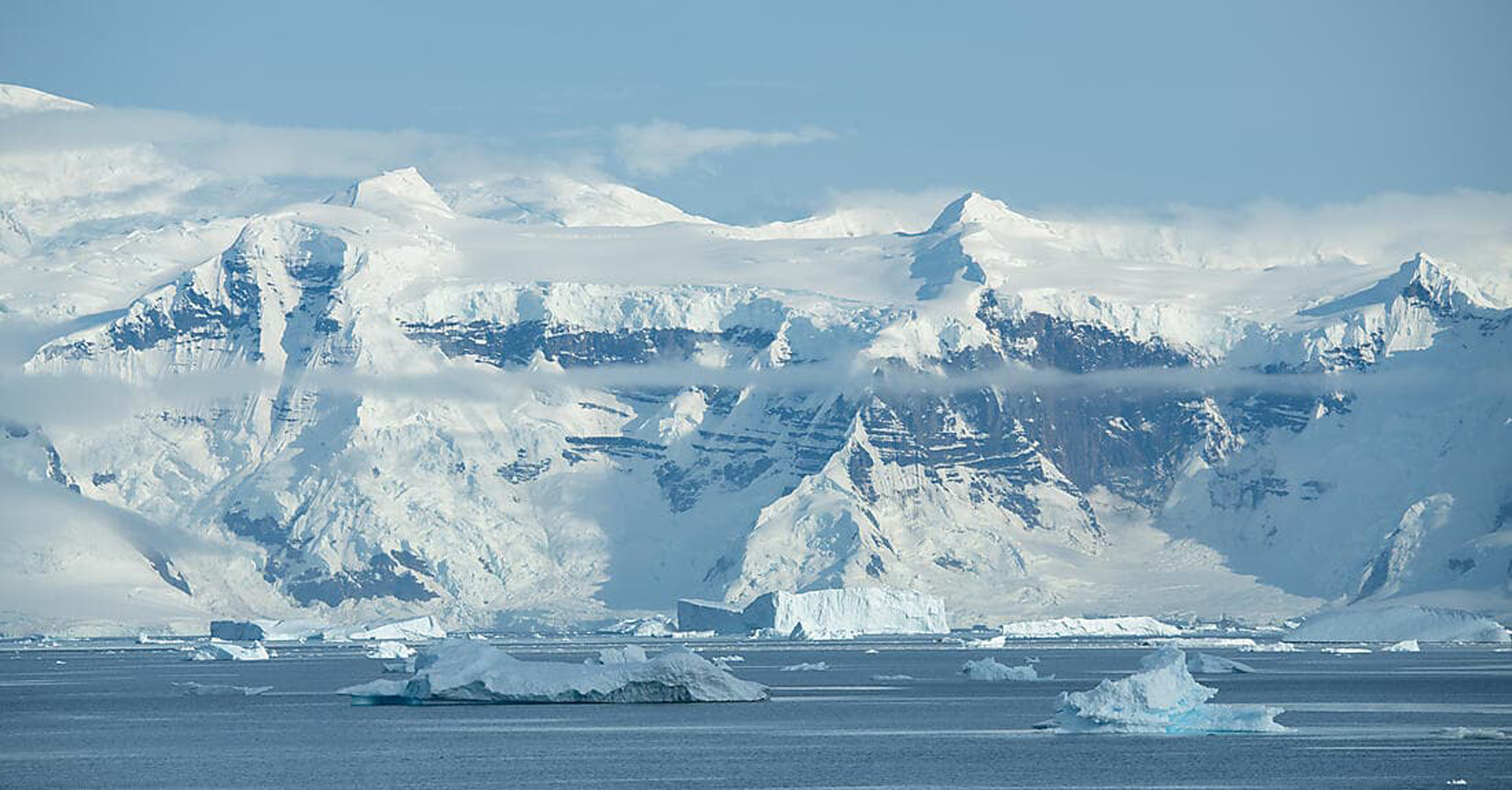 Snow-Capped Mountains and Icebergs in Antarctica A stunning polar landscape featuring towering snow-covered mountains rising dramatically from the sea, with numerous icebergs floating in the foreground. The pristine white peaks are partially shrouded in mist, creating a serene and majestic Antarctic seascape under clear blue skies.