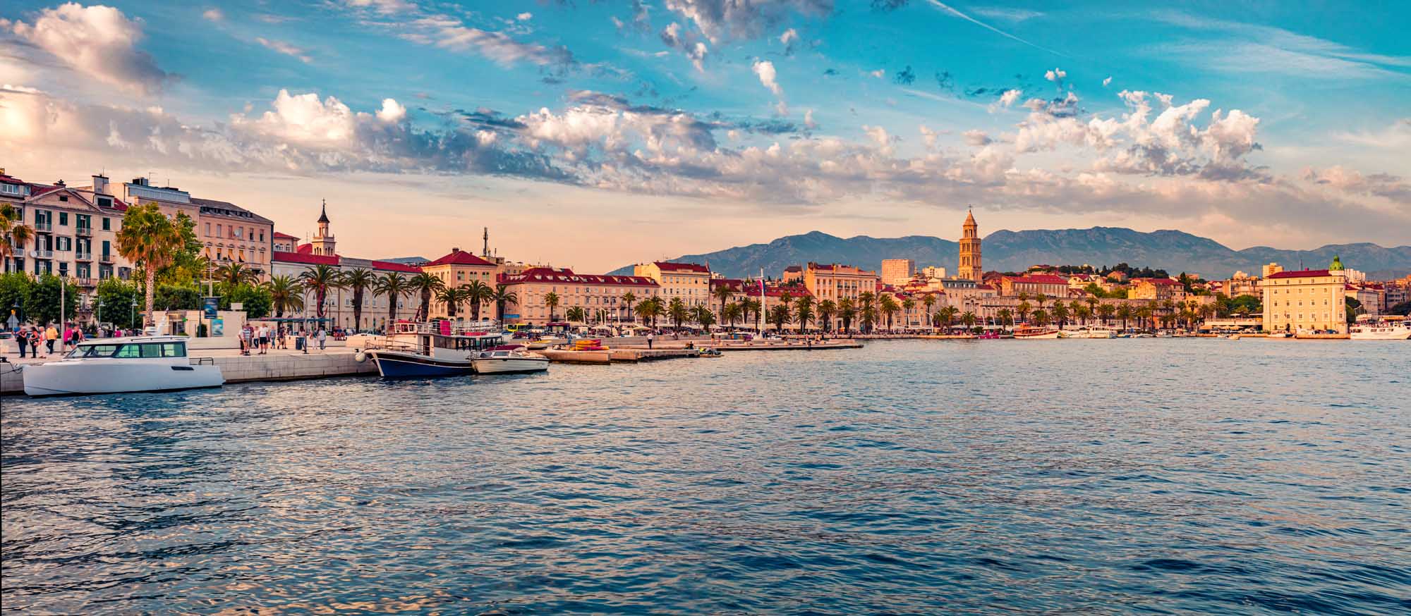 Split Waterfront at Golden Hour A stunning panoramic view of Split, Croatia's historic waterfront at sunset, featuring traditional Mediterranean architecture, palm-lined promenade, moored boats, and mountains in the distance under a dramatic blue sky with white clouds.