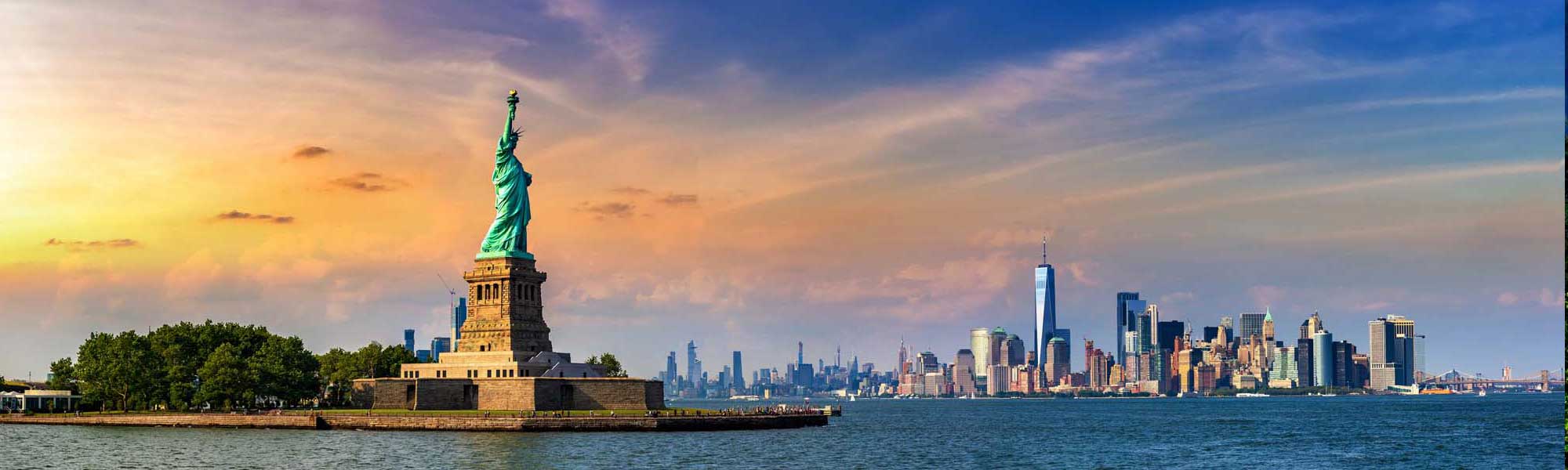 Statue of Liberty and Manhattan Skyline at Sunset An iconic view of the Statue of Liberty on Liberty Island with the Manhattan skyline illuminated in golden sunlight during sunset. The image captures the copper statue against a vibrant sky with the modern skyscrapers of lower Manhattan visible across the water.