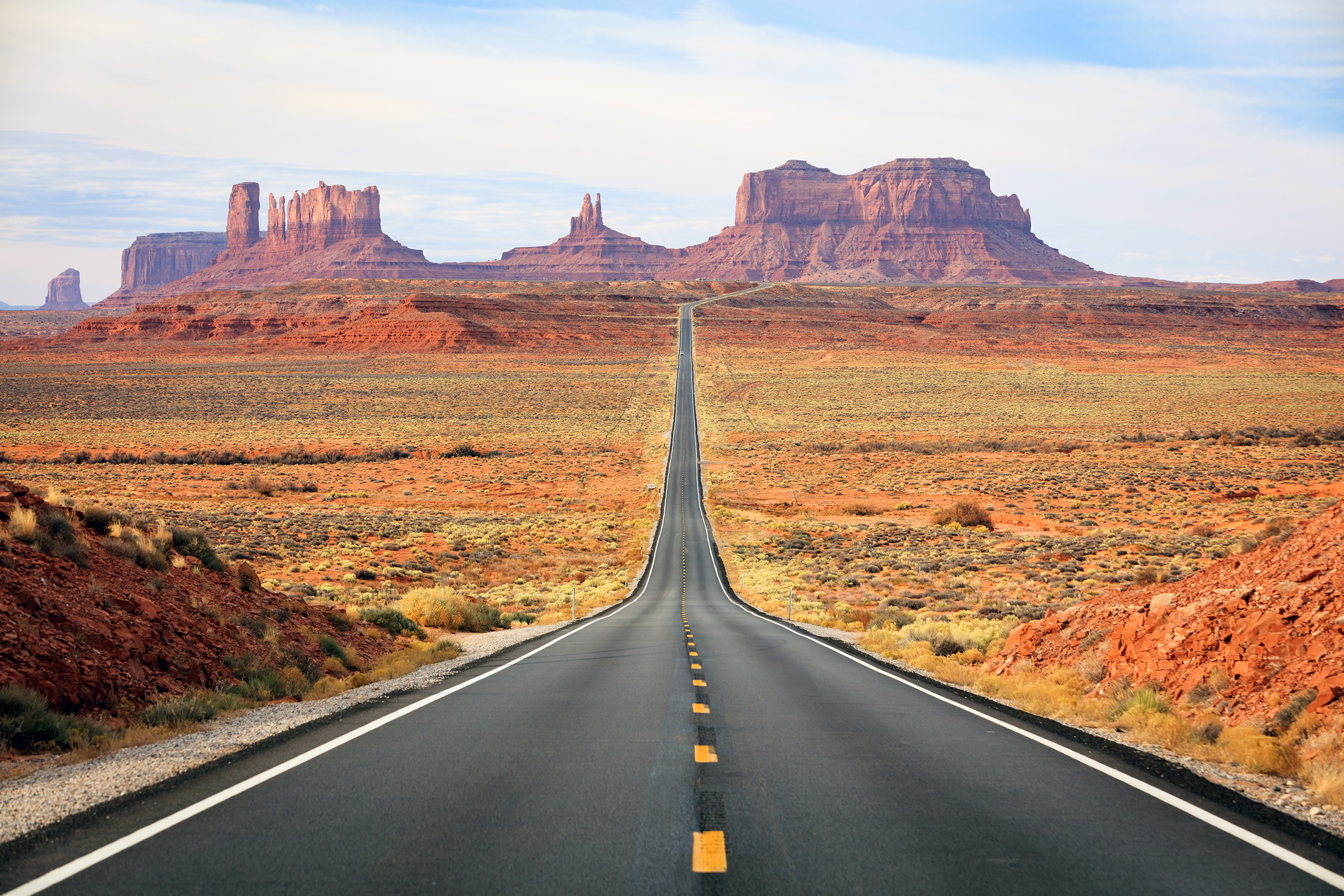 Straight Desert Highway to Monument Valley A perfectly straight asphalt highway stretches toward the horizon, leading directly to the iconic buttes and mesas of Monument Valley. The image captures the classic American Southwest landscape with striking red rock formations, desert scrubland, and a clear blue sky, exemplifying the scenic road trip experience.