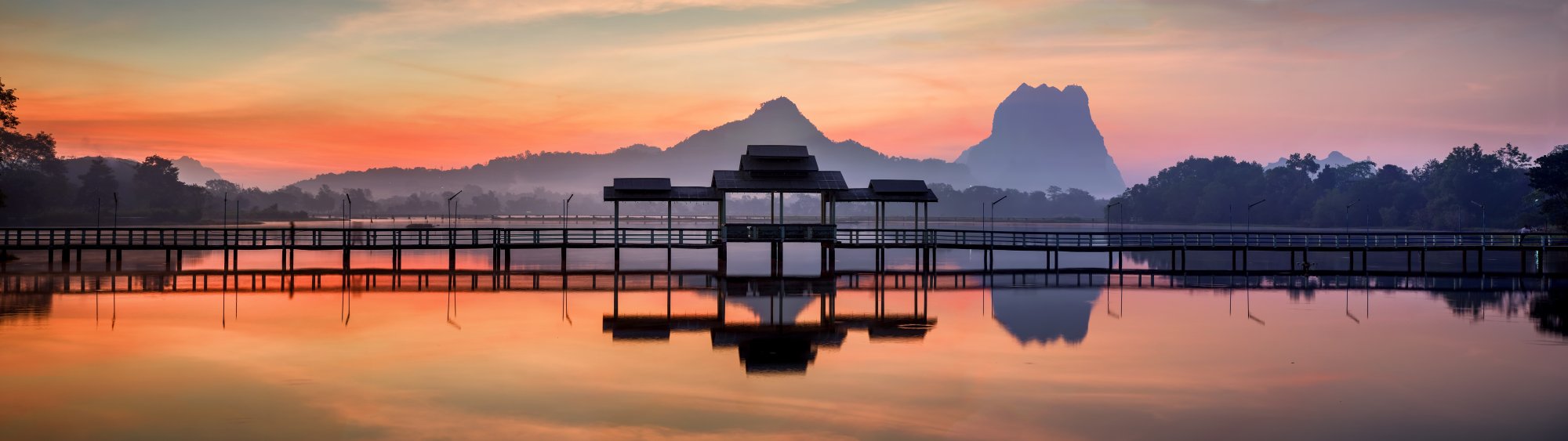 Sunrise Over Tropical Lake with Traditional Pavilion A serene sunrise scene over a calm lake featuring a traditional wooden pavilion structure with distinctive tiered roofing, silhouetted against dramatic mountain peaks in the background. The tranquil water perfectly mirrors the vibrant orange and pink sky, creating a symmetrical composition that evokes peaceful Southeast Asian landscapes.