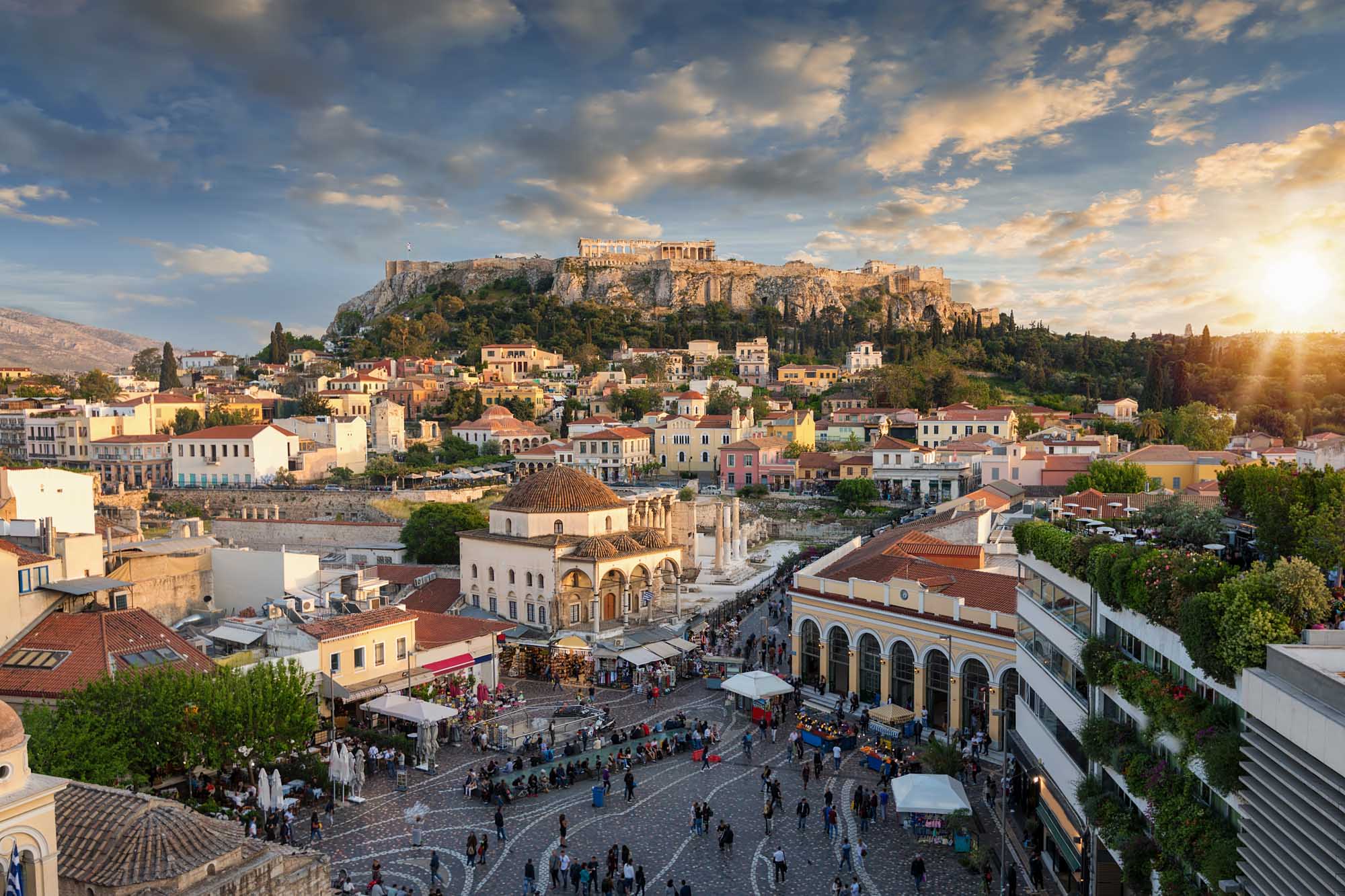 A stunning golden-hour view of Athens' historic Plaka district with colorful neoclassical buildings clustered below the illuminated Acropolis and Parthenon Temple perched atop a rocky hill. The scene captures the Byzantine Church of the Holy Apostles in the foreground with tourists gathered in the bustling square, exemplifying the blend of ancient history and vibrant modern life in Greece's capital.
