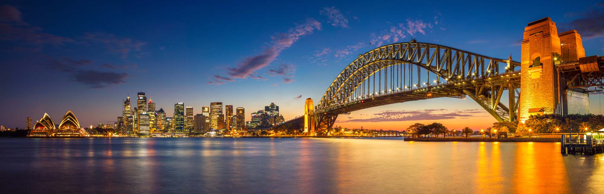 Sydney Harbour at Twilight with Opera House A stunning panoramic view of Sydney, Australia during the blue hour twilight, featuring the iconic Sydney Harbour Bridge illuminated in golden light, the distinctive Sydney Opera House on the left, and the glittering skyline reflected in the calm harbour waters.