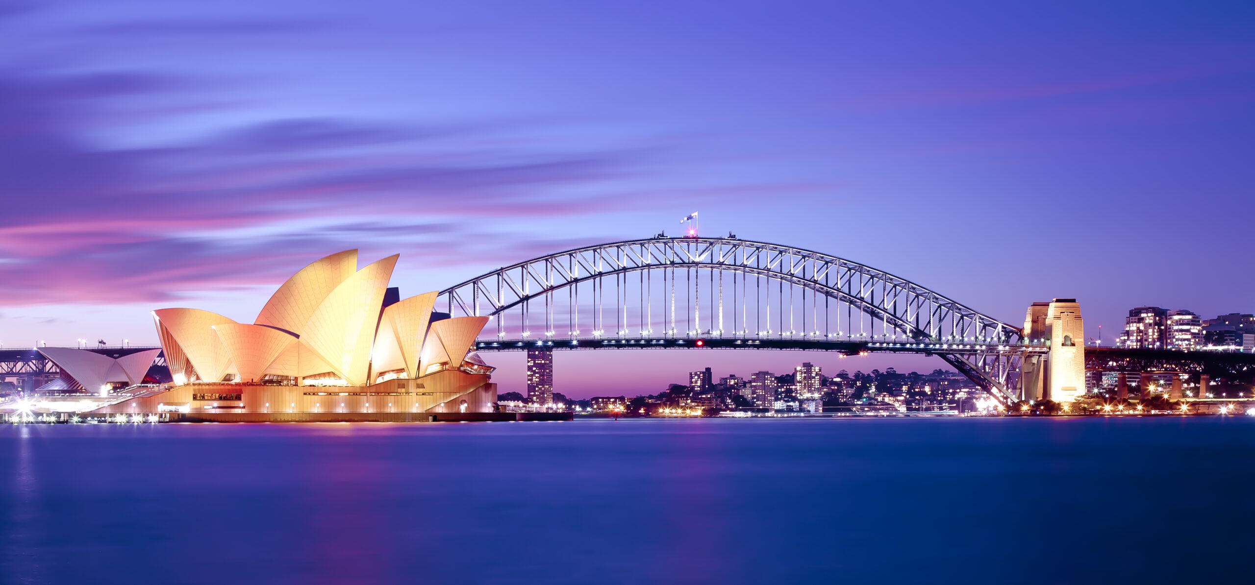 Sydney Opera House and Harbour Bridge at Twilight An iconic twilight photograph of Sydney's most famous landmarks, featuring the illuminated Sydney Opera House in the foreground and the Sydney Harbour Bridge spanning across the harbor against a stunning purple and pink sunset sky, with the city's skyline lights twinkling in the background.