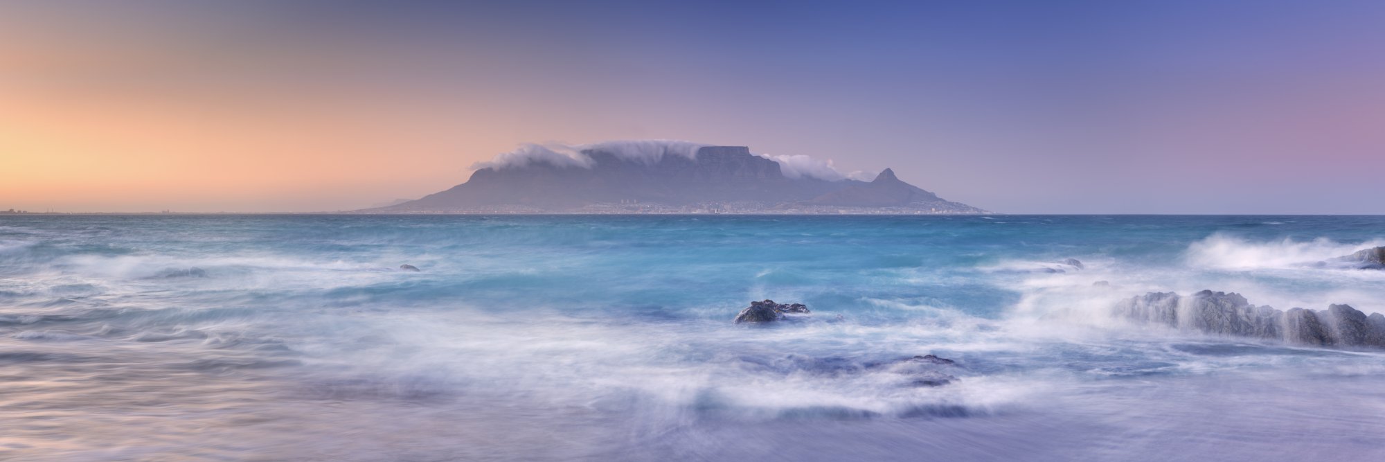 Table Mountain at Sunrise from Beach A stunning coastal landscape featuring Table Mountain shrouded in cloud cover at dawn, viewed across turquoise waters with rolling waves and rocky outcrops in the foreground. The image captures the iconic flat-topped mountain of Cape Town during golden hour, with soft purple and blue tones dominating the sky.