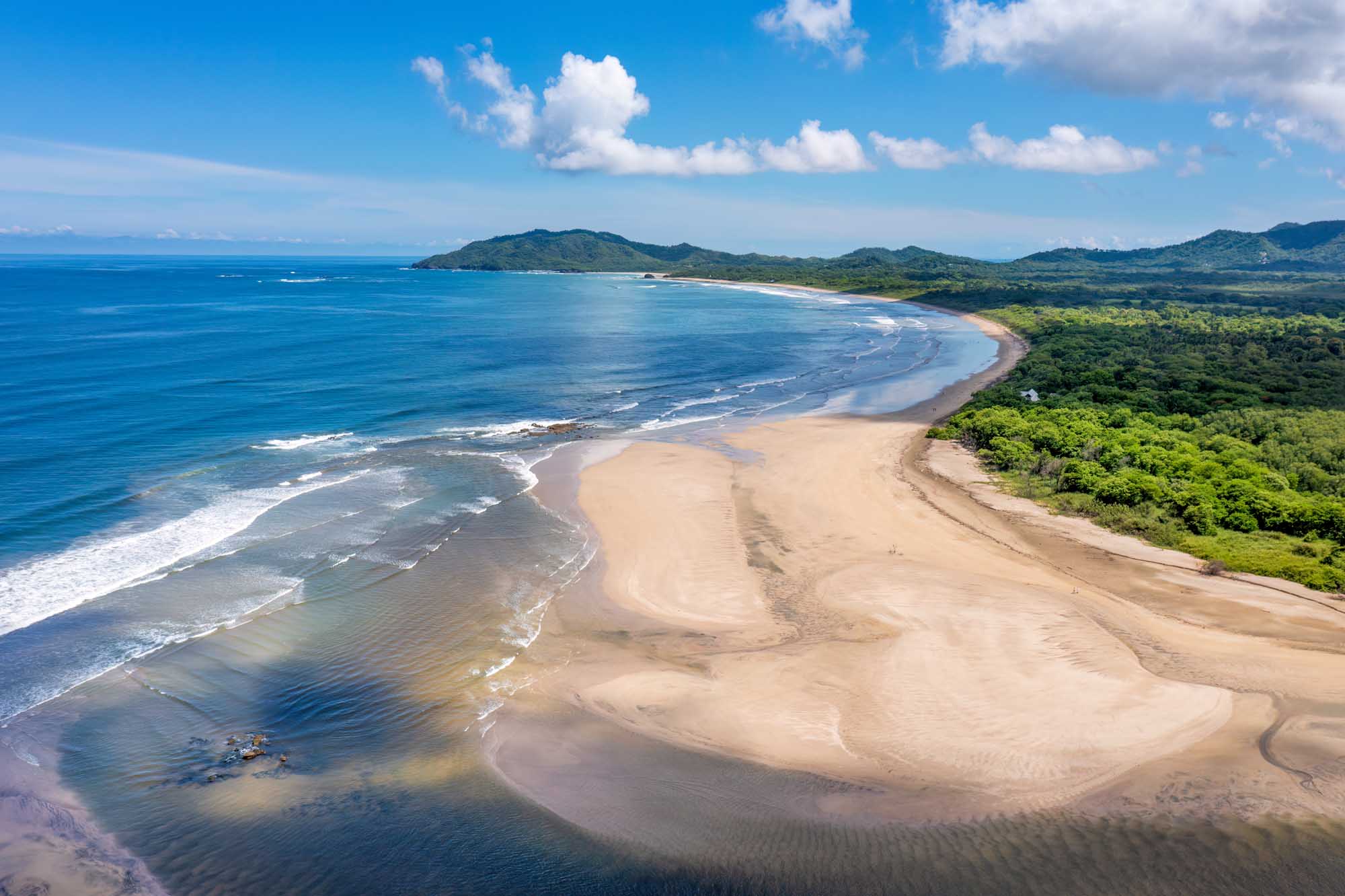 An aerial view of Tamarindo Beach and the Tamarindo River estuary in Guanacaste, Costa Rica, showing the pristine sandy beach curving along the Nicoya Peninsula with lush tropical vegetation, river mouth, and turquoise Pacific waters with white-capped waves under a bright blue sky.