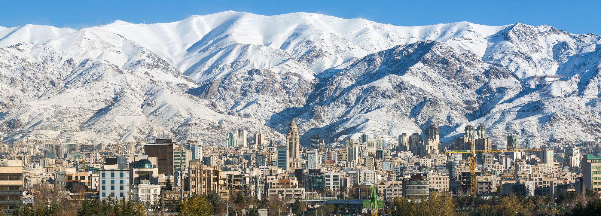 Tehran Skyline with Snow-Capped Alborz Mountains A stunning urban landscape showing the dense cityscape of Tehran, Iran, dominated by numerous high-rise buildings and traditional architecture, set against the dramatic backdrop of the snow-covered Alborz mountain range under a clear blue sky.