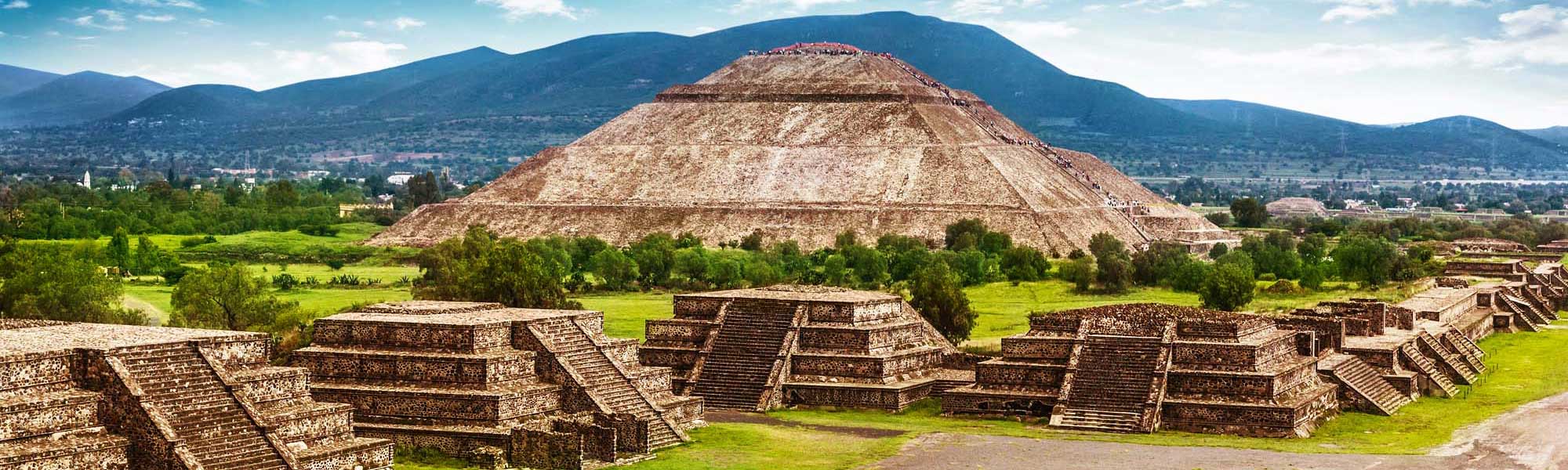 The iconic Pyramid of the Sun dominates this expansive view of Teotihuacan's archaeological complex in Mexico, with smaller pyramidal structures and ancient foundations scattered across verdant grounds, set against blue mountains and a clear sky.