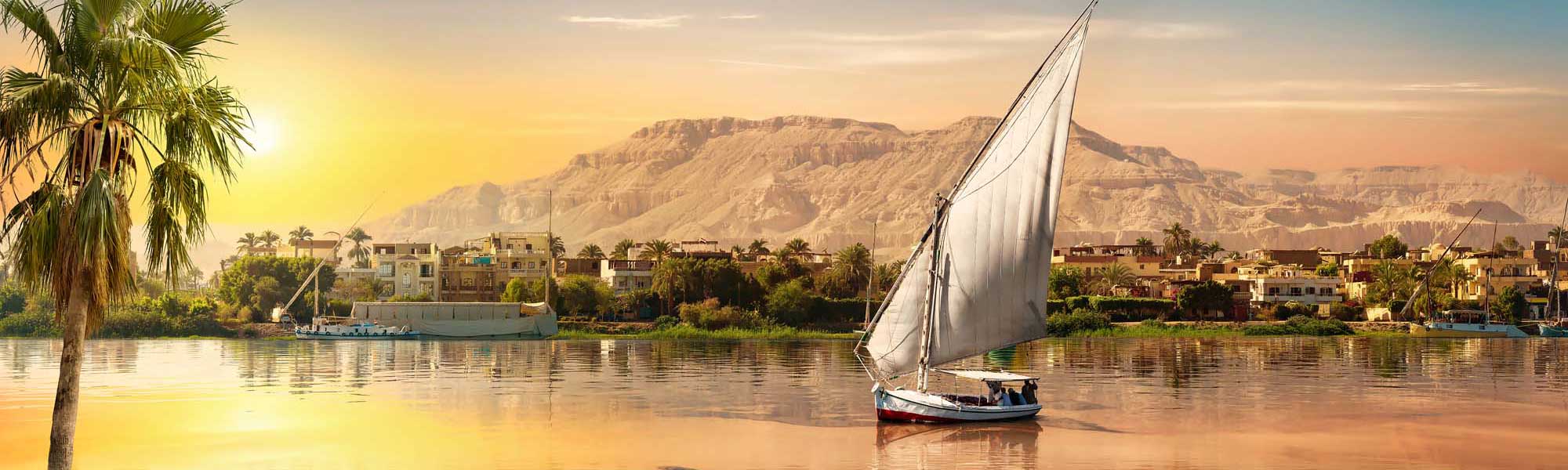 A traditional Egyptian felucca sailboat with white canvas sails glides peacefully across the Nile River at golden sunrise, with palm-lined riverbanks, colorful buildings, and desert cliffs in the background. The warm morning light bathes the scene in golden and amber tones, capturing the timeless beauty of river travel in Egypt.