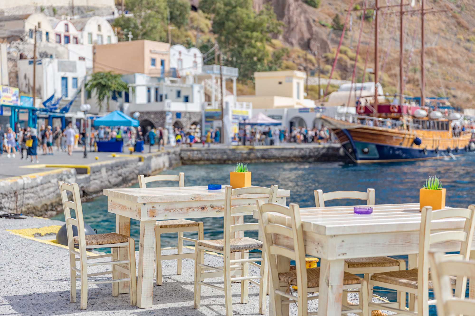 A charming waterfront taverna in Santorini, Greece, featuring whitewashed wooden tables and chairs with woven rush seats overlooking a bustling harbor. The scene captures traditional Cycladic architecture with blue-trimmed buildings, moored sailing vessels, and tourists exploring the picturesque port village.