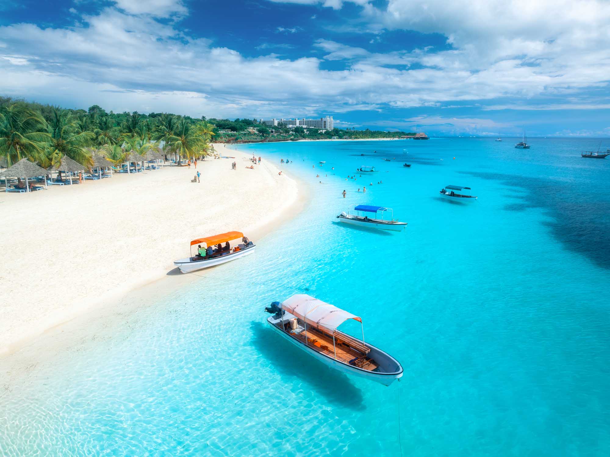 Tropical Beach with Moored Boats at Kendwa, Zanzibar An aerial drone view of a pristine tropical beach featuring crystal-clear turquoise waters, white sand, traditional fishing boats moored offshore, palm trees lining the shore, and a beachfront resort in the background under a bright blue sky.