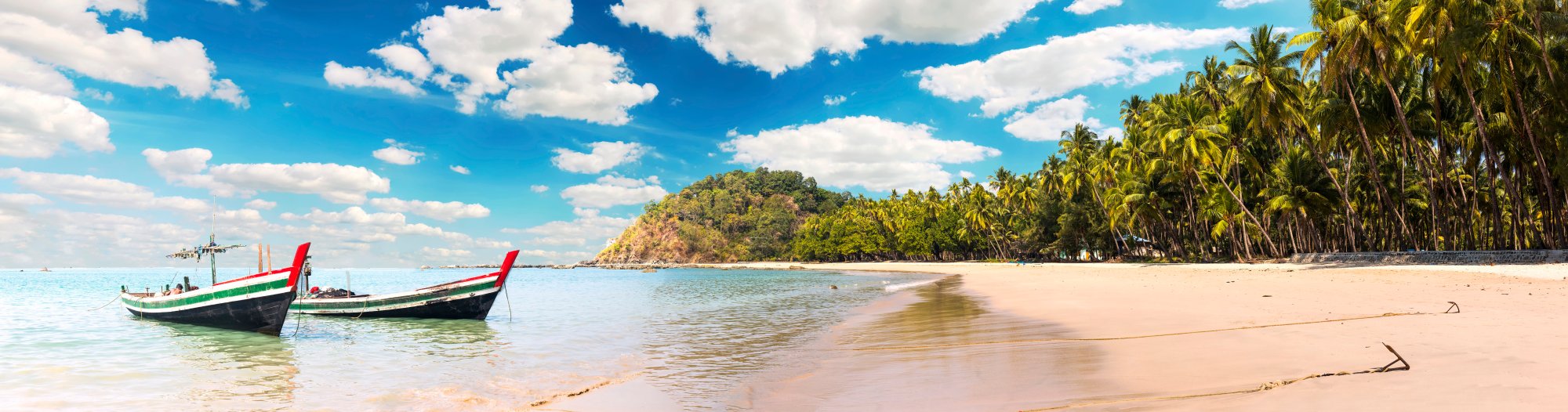Tropical Beach with Traditional Long-Tail Boats A picturesque tropical beach scene featuring two traditional colorful long-tail boats anchored in crystal-clear turquoise waters, with pristine white sand, lush palm trees, and a forested hillside under a brilliant blue sky with white clouds.