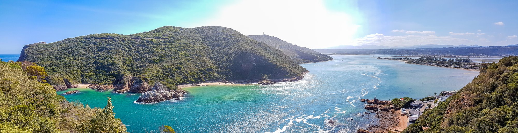 Tropical Coastal Bay with Forested Headland A stunning panoramic view of a pristine tropical coastline featuring a verdant, densely forested headland jutting into crystal-clear turquoise waters. The scene showcases a secluded sandy beach nestled in a protected cove, with distant settlements visible across the bay and calm blue waters extending to the horizon under clear skies.