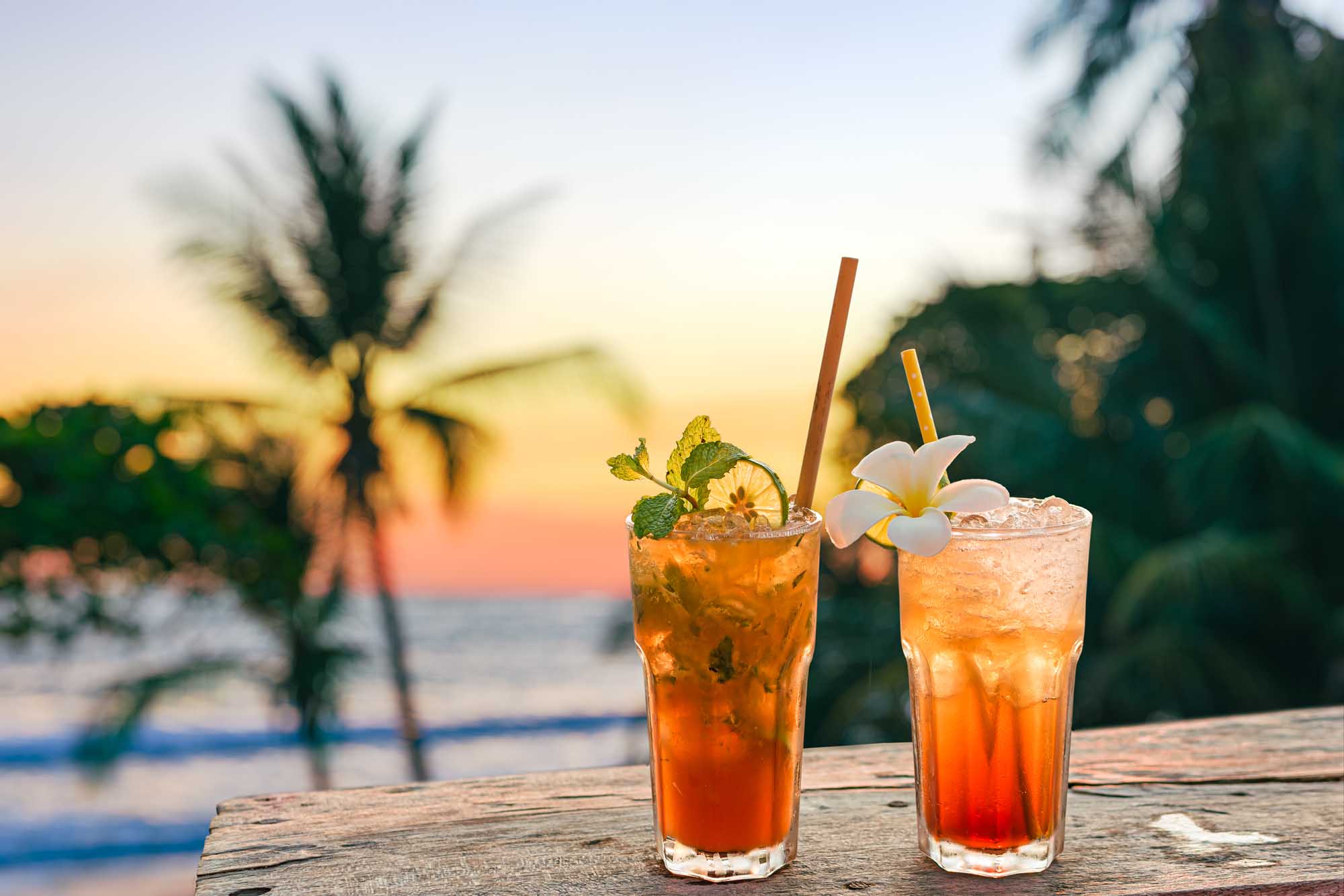 Two vibrant tropical cocktails with orange and amber hues sit on a weathered wooden table overlooking a beach at sunset. The drinks are garnished with fresh mint, lime slices, and white flowers, with palm trees and ocean waves visible in the soft-focus background during golden hour.