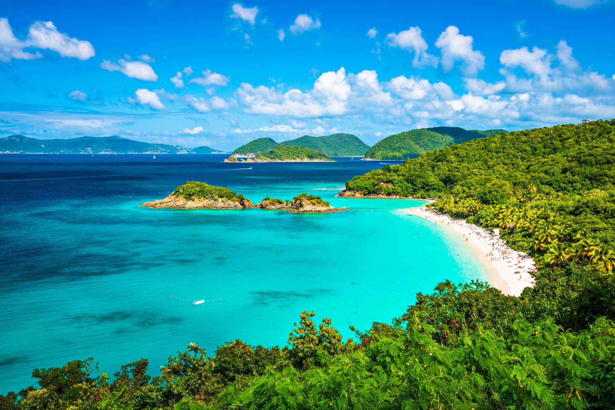 A breathtaking tropical paradise featuring pristine turquoise waters, white sandy beaches, and lush green vegetation. Small rocky islands dot the crystalline bay while sailboats anchor in the calm water, with verdant hillsides and distant islands framing the spectacular seascape under a brilliant blue sky.