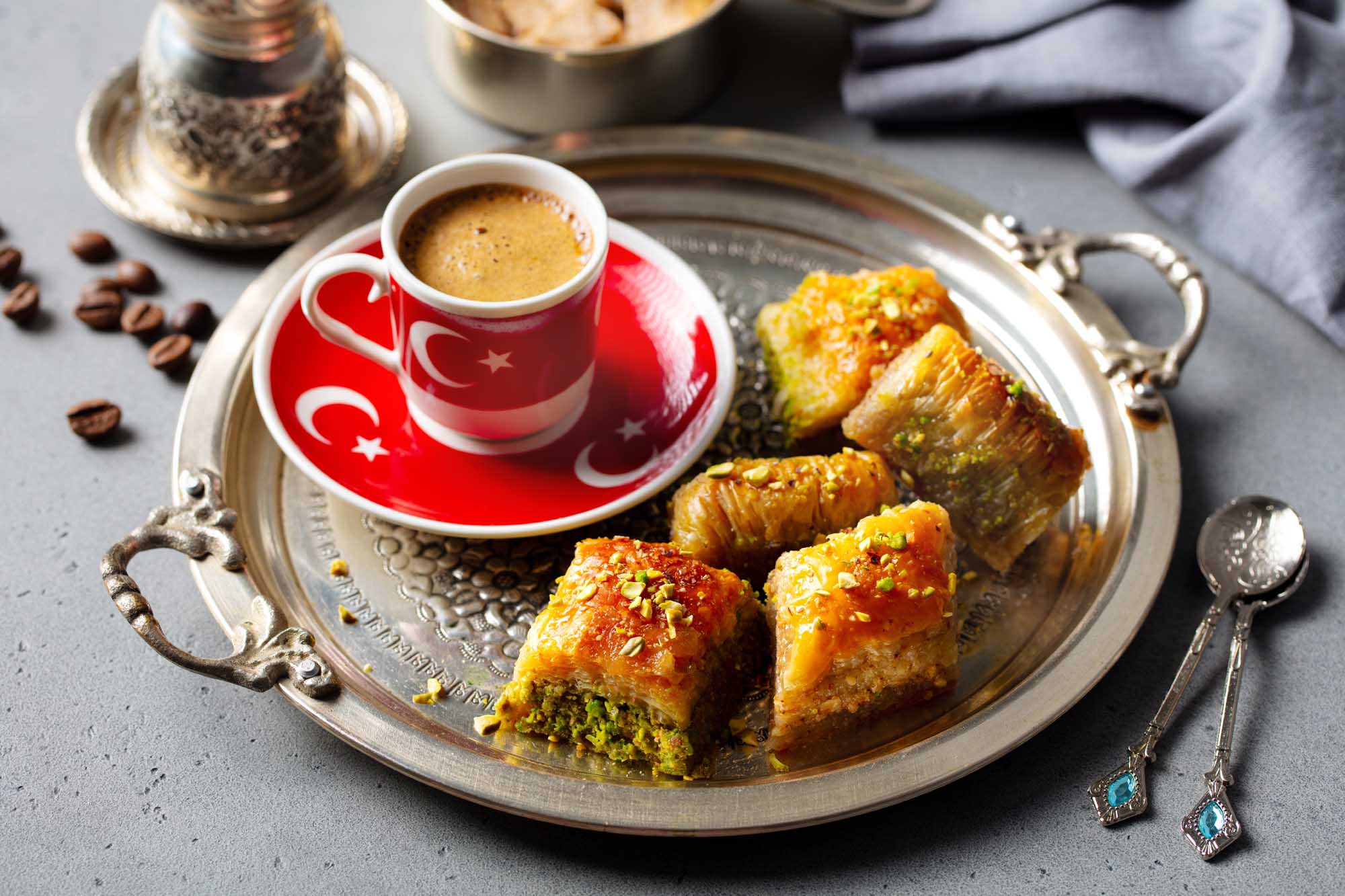 An elegant presentation of traditional Turkish coffee served in a red cup with Turkish flag motifs, accompanied by golden baklava pastries topped with pistachios on an ornate silver tray. The composition includes scattered coffee beans, decorative silver utensils, and traditional Turkish coffee serving accessories, showcasing authentic Middle Eastern culinary culture.