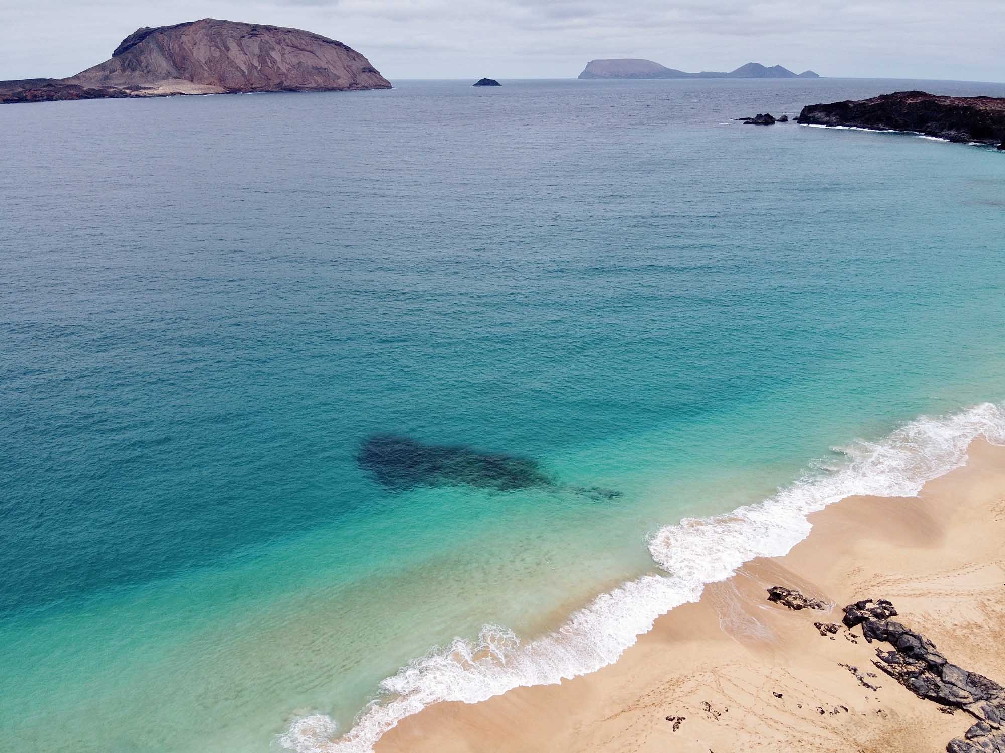 A pristine sandy beach with crystal-clear turquoise waters and gentle white-capped waves, featuring dramatic volcanic islands visible in the distance across a calm sea. The landscape showcases volcanic rock formations along the shoreline and a distinctive cone-shaped island on the left horizon.