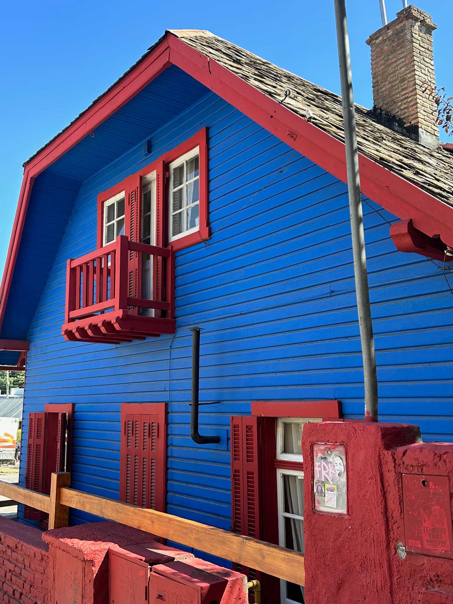 Vibrant Blue and Red Beachside House A striking residential building painted in bold blue with bright red trim and accents, featuring a distinctive pitched roof and upper-level balcony. The charming cottage-style architecture showcases red louvered shutters, red doors at ground level, and a brick chimney, exemplifying colorful coastal architectural traditions.