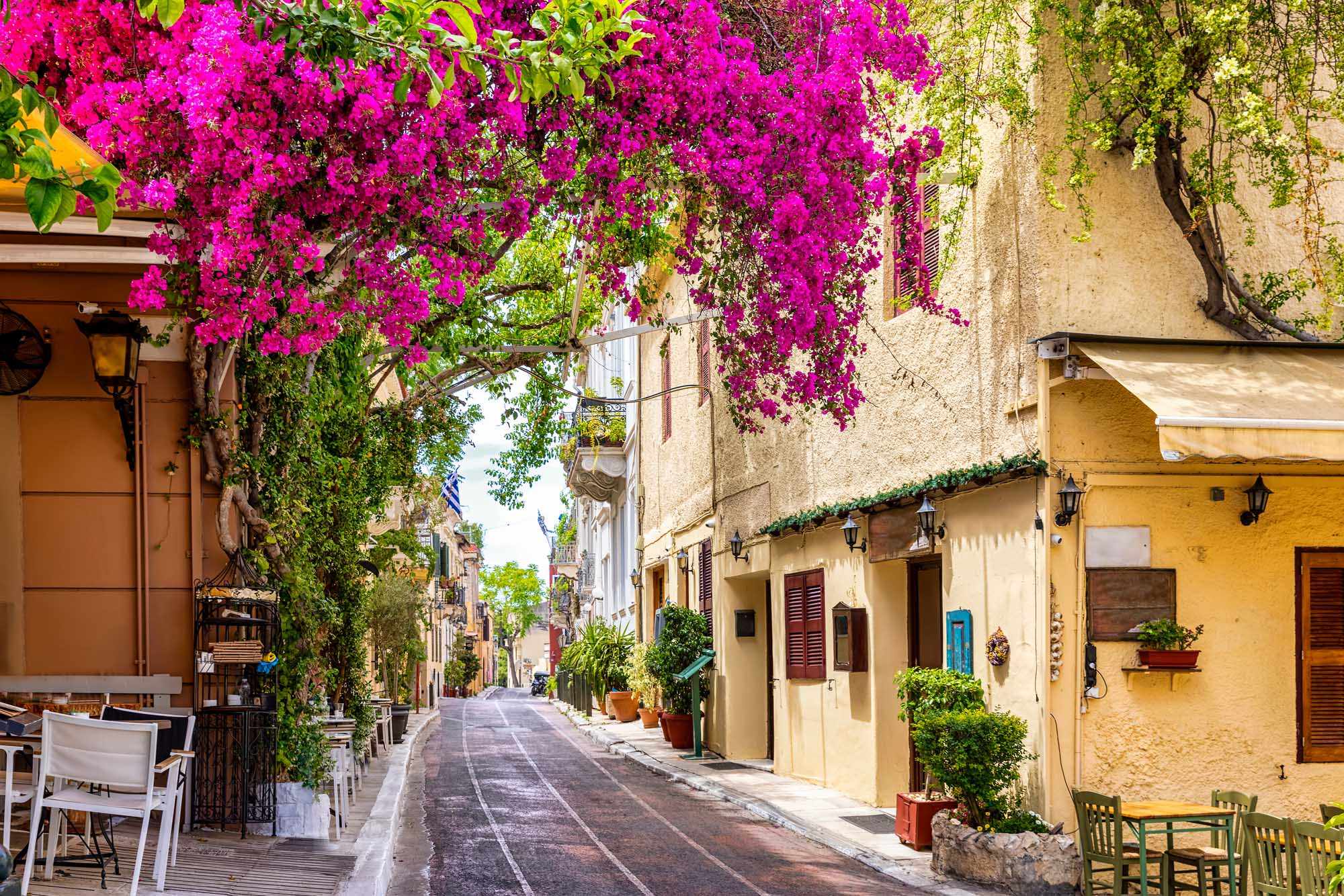 A charming Mediterranean alleyway lined with traditional stone buildings and cascading magenta bougainvillea flowers overhead, featuring characteristic architecture with colorful doors, shutters, and outdoor café seating along a quiet street.
