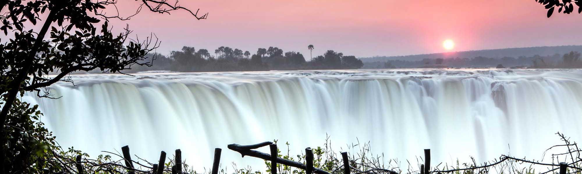 A breathtaking view of Victoria Falls at sunrise, with massive cascading water creating a luminous white curtain against a pink dawn sky. The iconic waterfall is framed by lush vegetation and trees along the Zambezi River, with the sun rising majestically in the background.