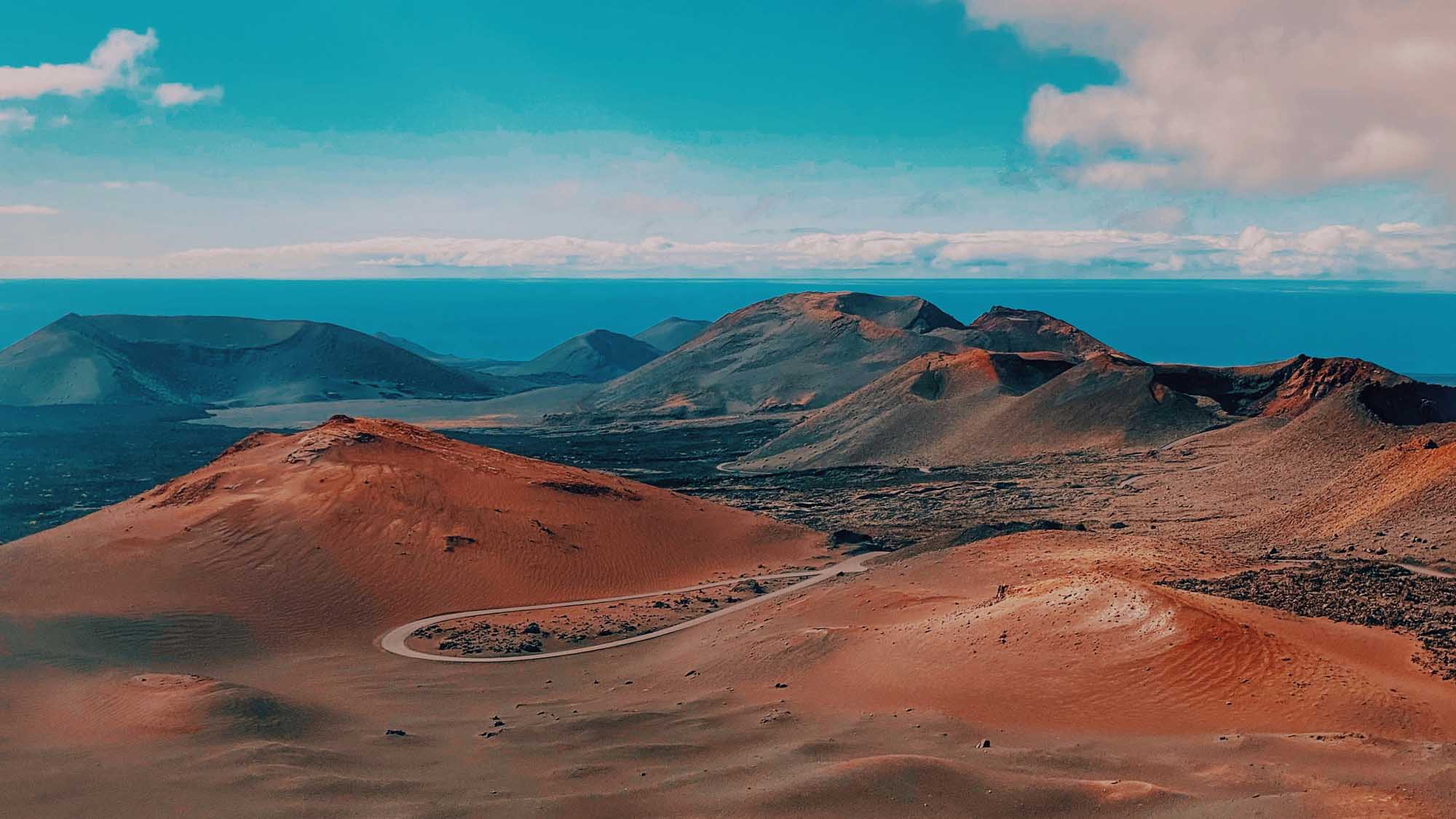 A dramatic volcanic landscape featuring distinctive rust-red and dark cinder cones rising above a vast lava field, with a striking turquoise ocean and distant islands visible on the horizon. The arid terrain displays the raw geological beauty of a volcanic archipelago with minimal vegetation.