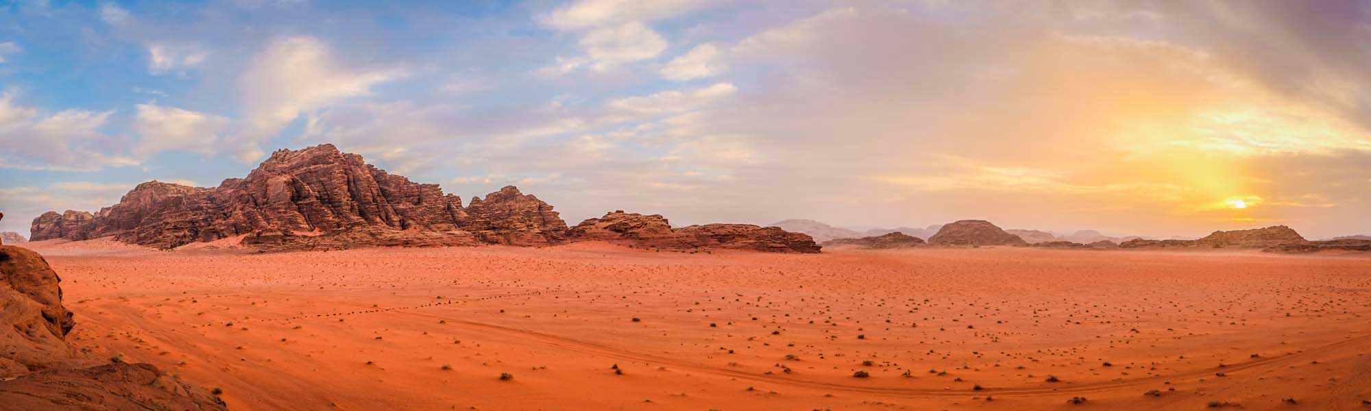 A breathtaking panoramic view of the Wadi Rum desert in Jordan, featuring towering rust-colored sandstone rock formations rising from the expansive red sand plains during golden hour, with a dramatic sky transitioning from blue to warm orange and yellow tones.